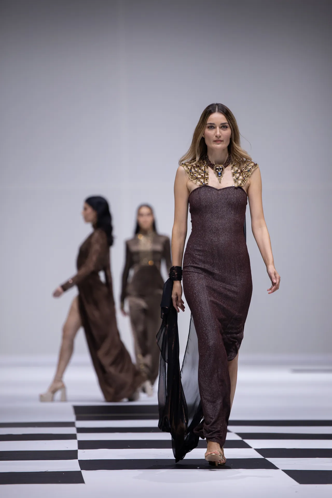 Model walking a runway in a burgundy sequined gown with a gold jeweled collar; black-and-white checkerboard floor underfoot.