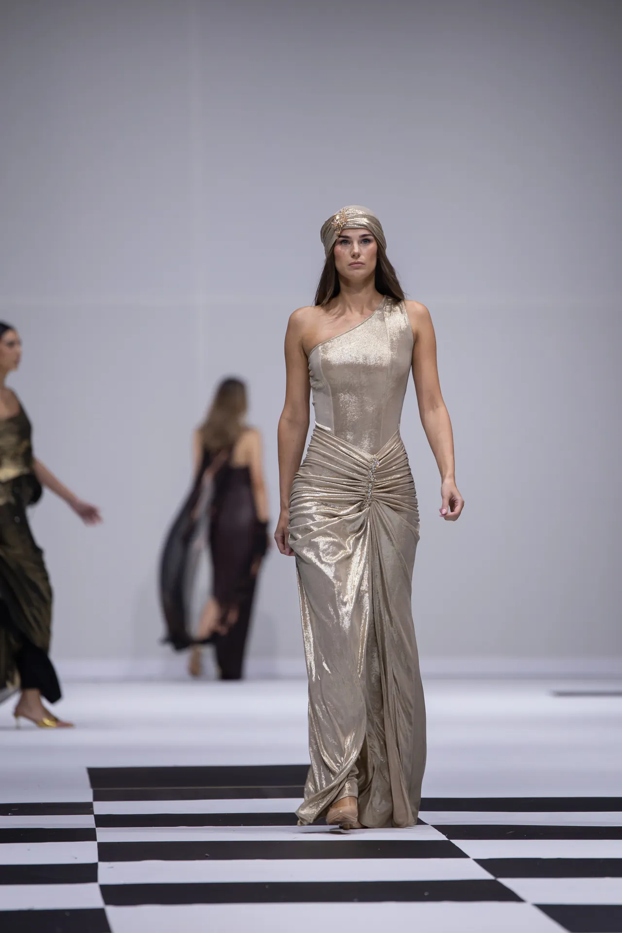 Model on a fashion runway wearing a metallic gold one-shoulder gown and matching headwrap, walking down a black-and-white checkered catwalk.
