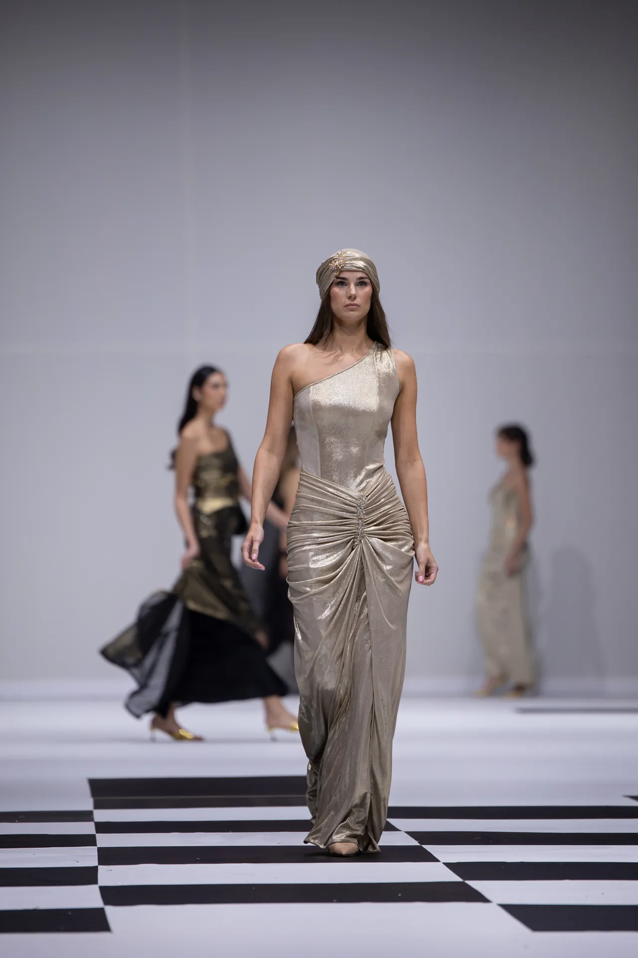 Model walks a shimmering gold one-shoulder gown down a black-and-white checkered runway with a matching head wrap.