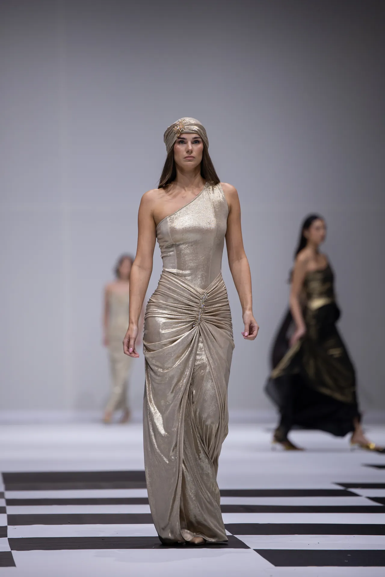 Model walking a metallic, one-shoulder gown with draped detailing and a matching head wrap on a black-and-white checkered runway.