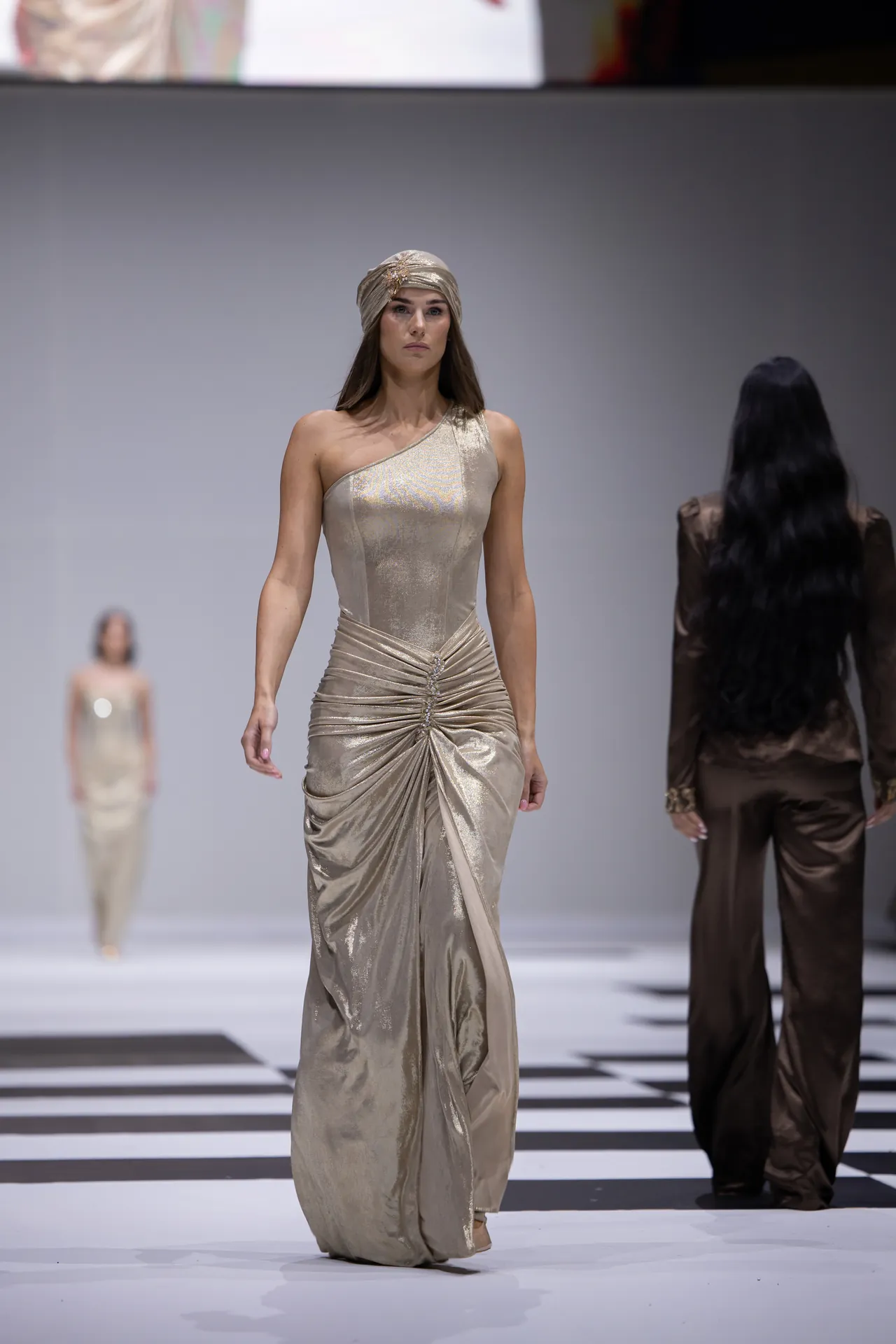 Model walks the runway in a metallic champagne one-shoulder gown with a matching turban headpiece, others visible in background.