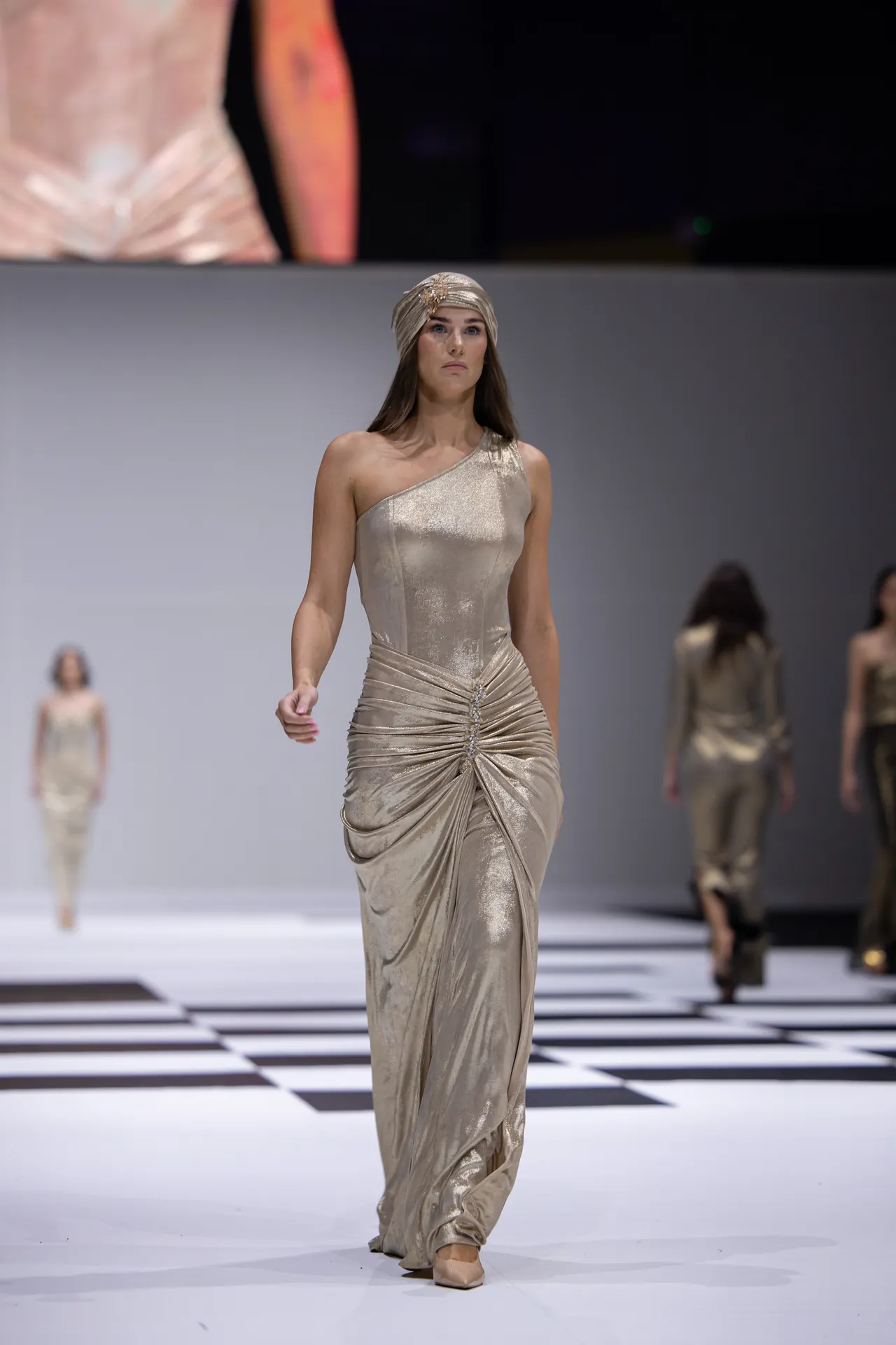 Model walks a metallic gold one-shoulder gown with a twisted draped center and matching headwrap on a fashion runway.