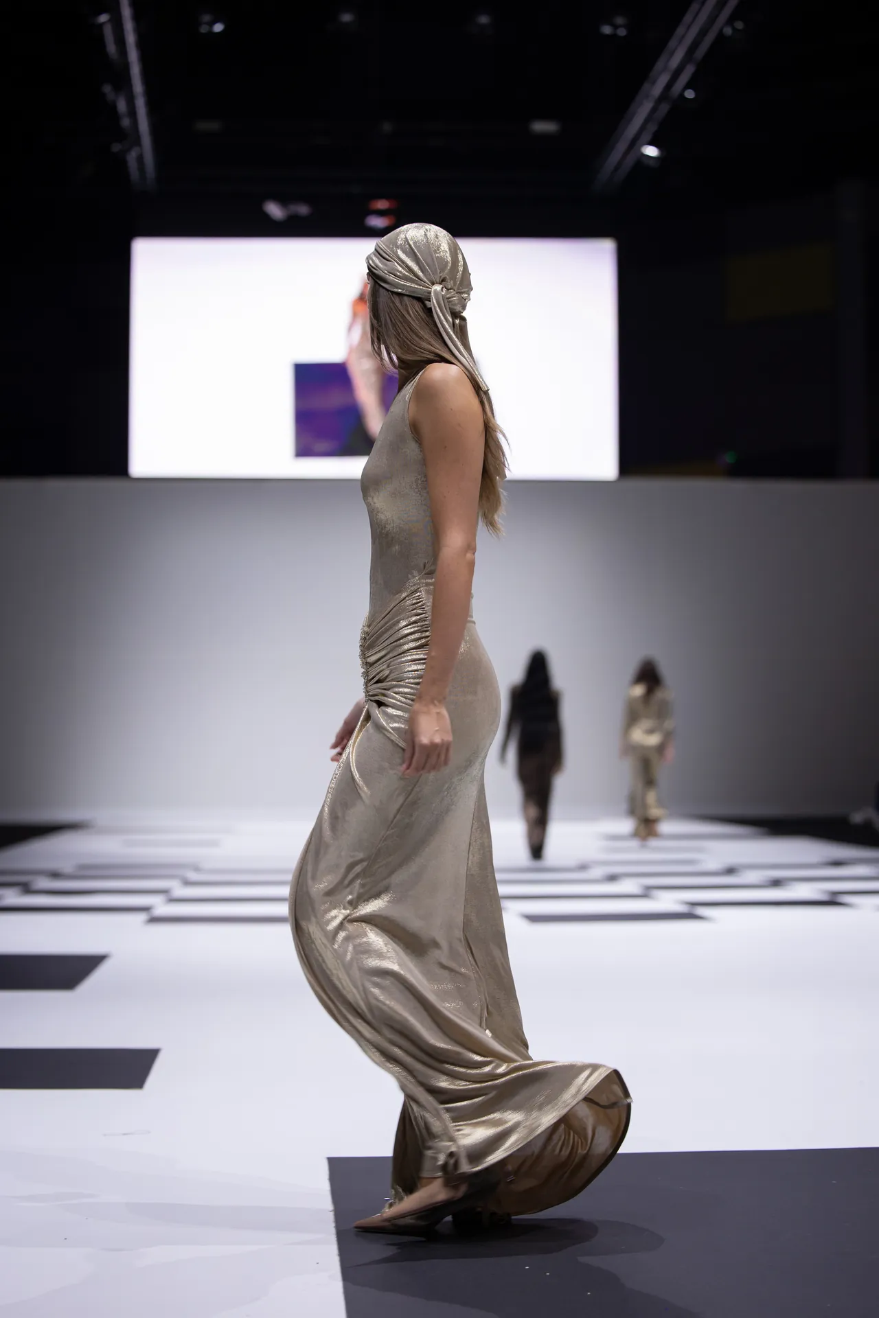 Model walking a metallic gold gown on a fashion runway with a matching headscarf, spotlight on the airy catwalk backdrop.
