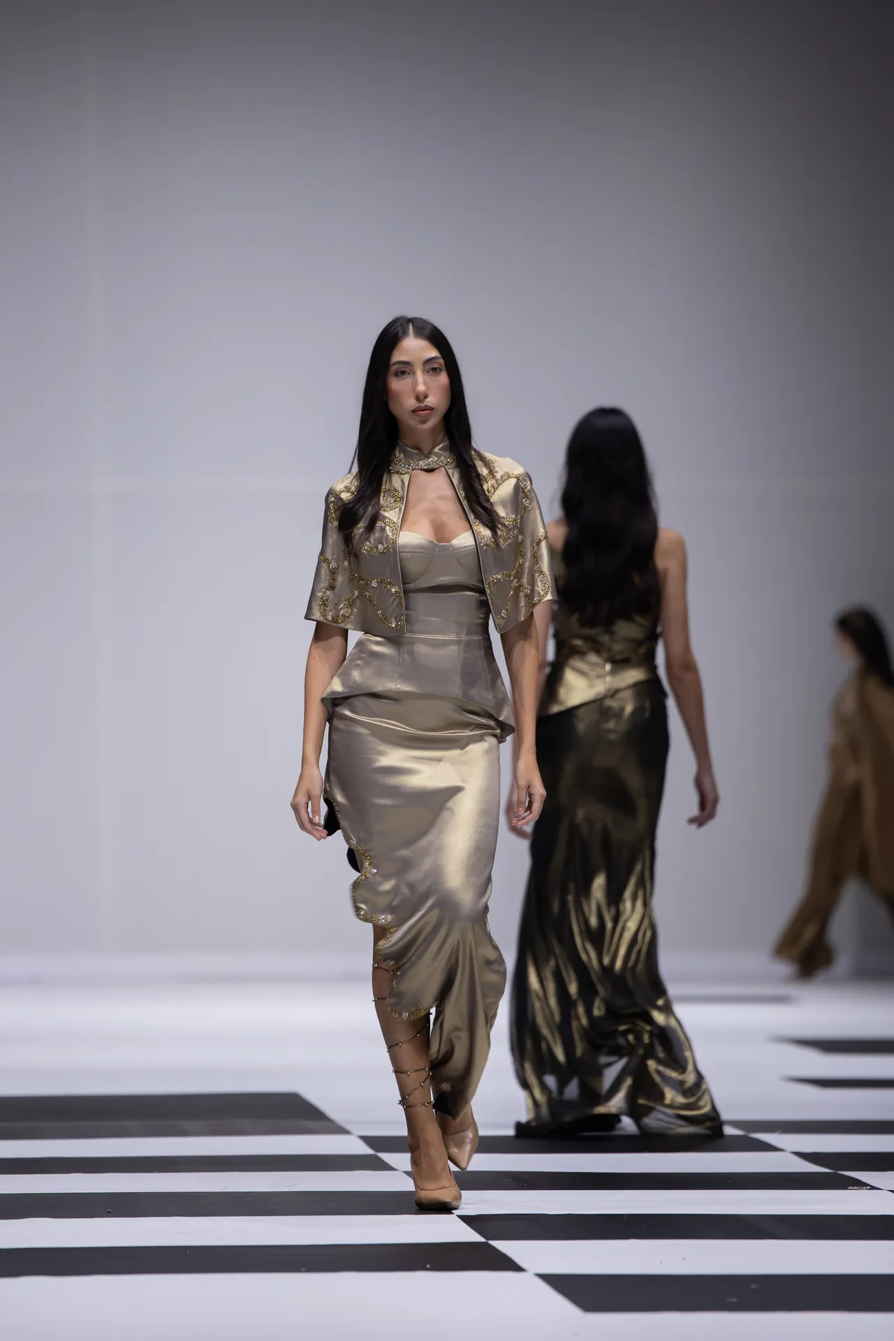 Model walks the runway in a metallic gold satin gown with an embroidered cape on a black-and-white checkered runway.