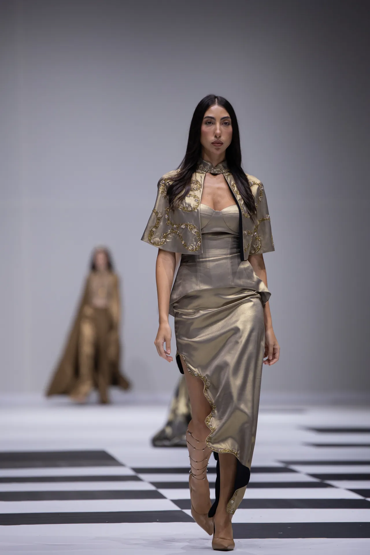 Model walks a metallic gold satin dress with ornate gold embroidery on the shoulders, on a black-and-white checker runway.