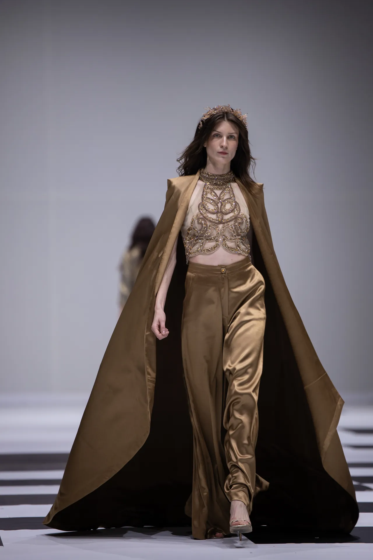 Model walking runway in metallic gold ensemble with long cape and intricate bodice, wearing a crown-like headpiece.