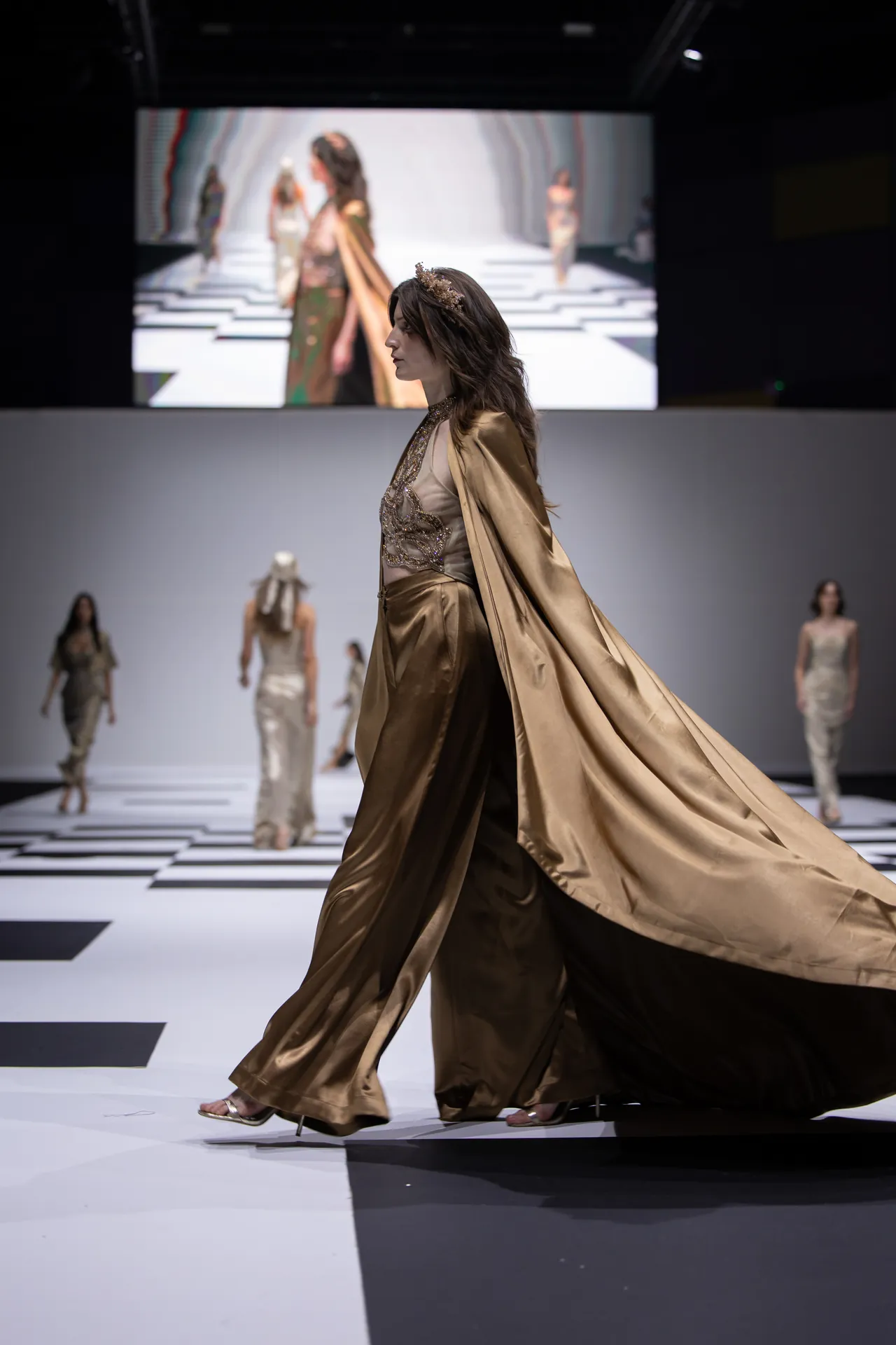 Model walking the runway in a gold satin gown with a long flowing cape and jeweled headpiece.