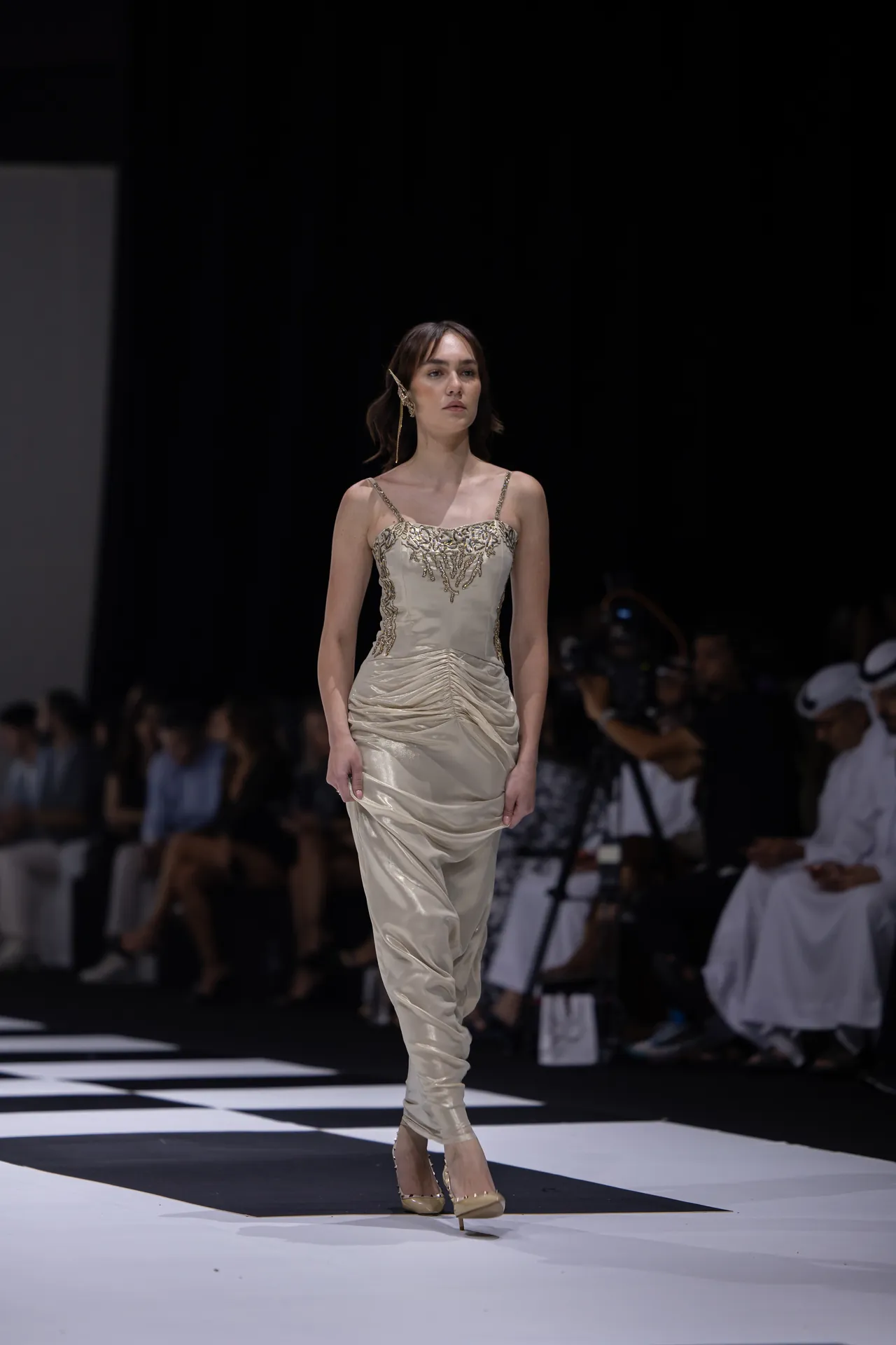 Model walking the runway in a cream satin gown with gold embroidery, with a blurred audience in the background.