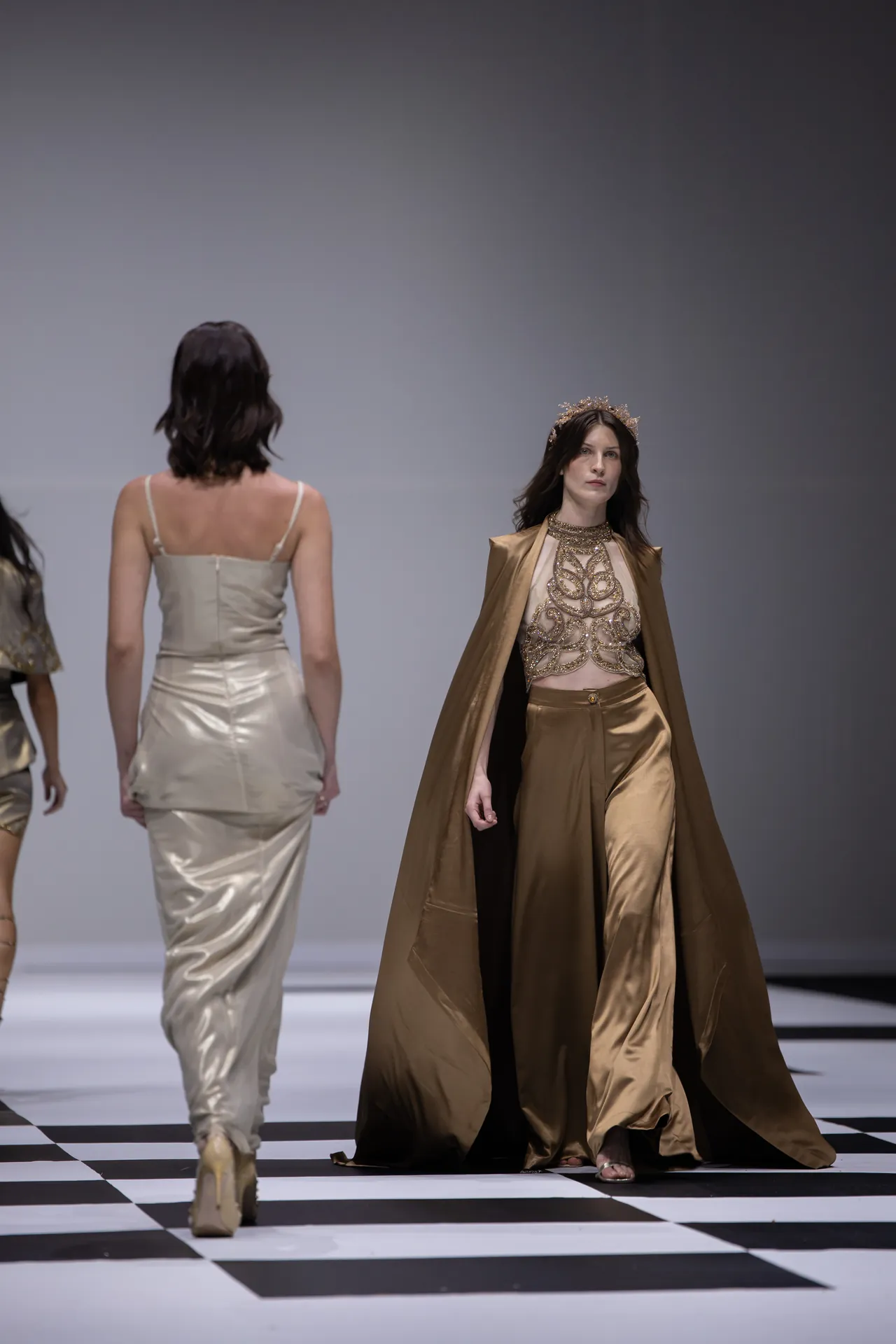 Model wearing a gold cape and intricately beaded top walks a fashion runway with a crown, while other beige satin gowns appear in the background on a checkered floor.