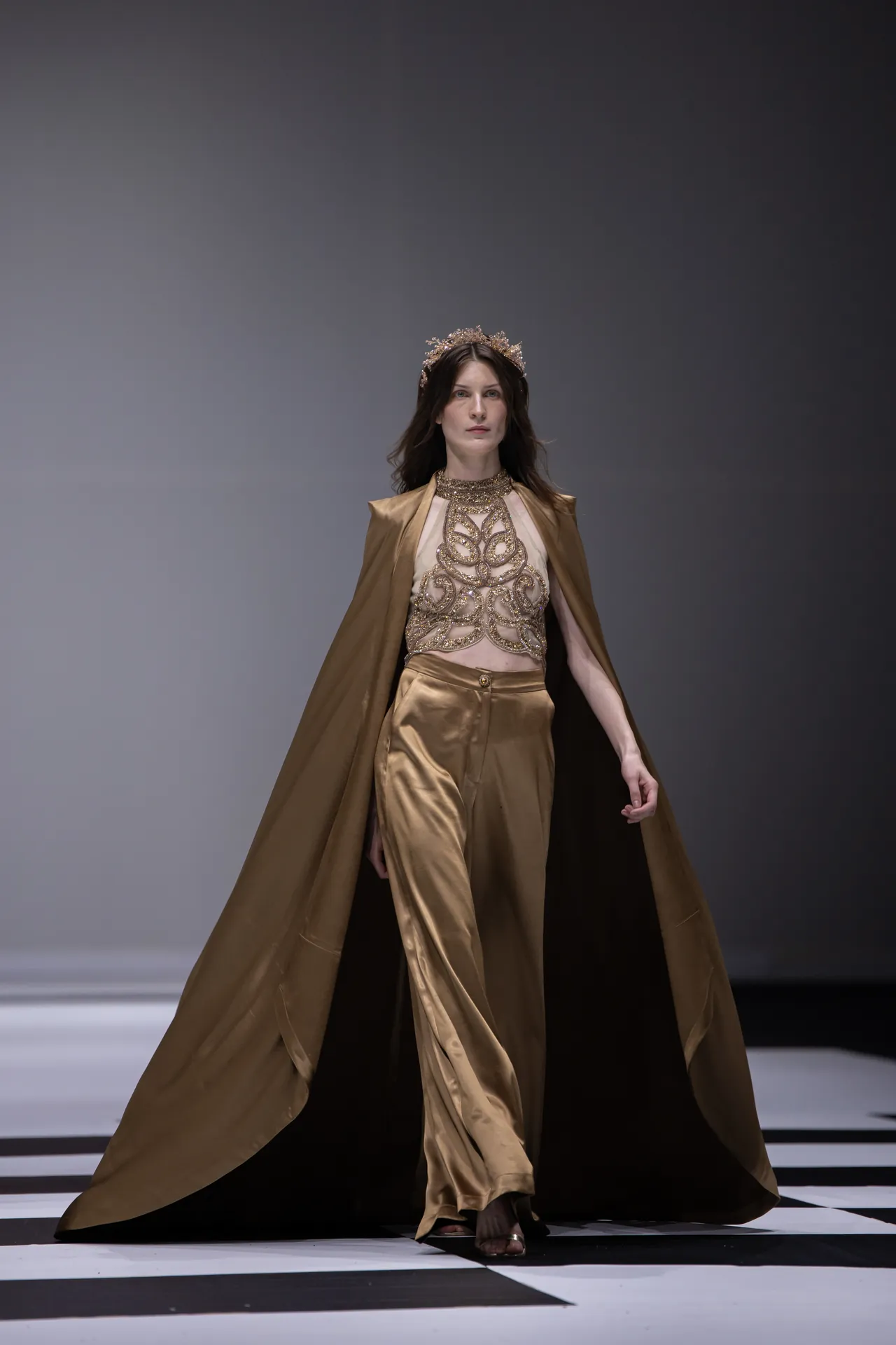 Model walking a fashion runway in a gold satin cape and embroidered top, wearing a decorative crown.