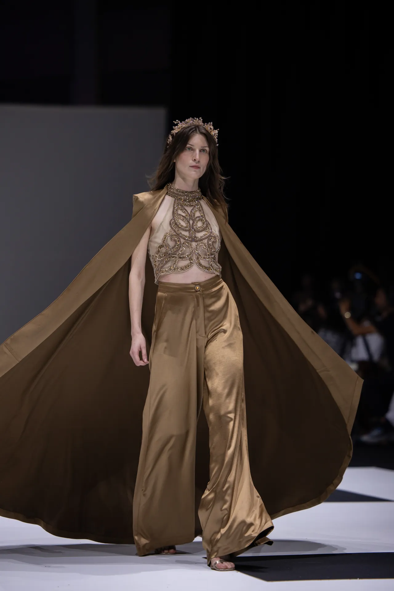 Model walks a fashion runway in a gold satin ensemble with embroidered bodice and a flowing cape, wearing a decorative gold crown.