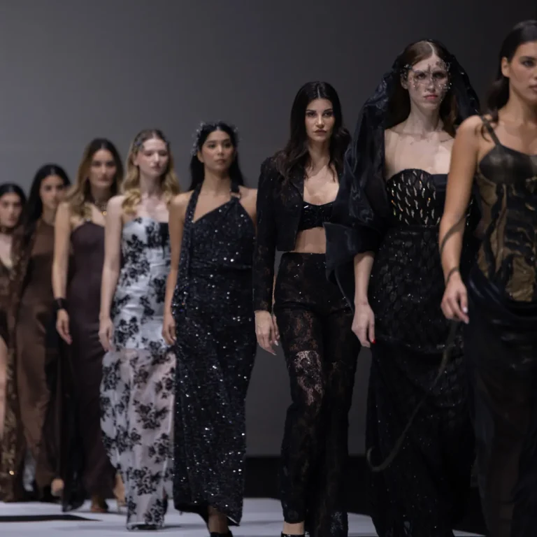 Models in dark, glittery outfits walk the fashion runway on a black-and-white checkered floor.