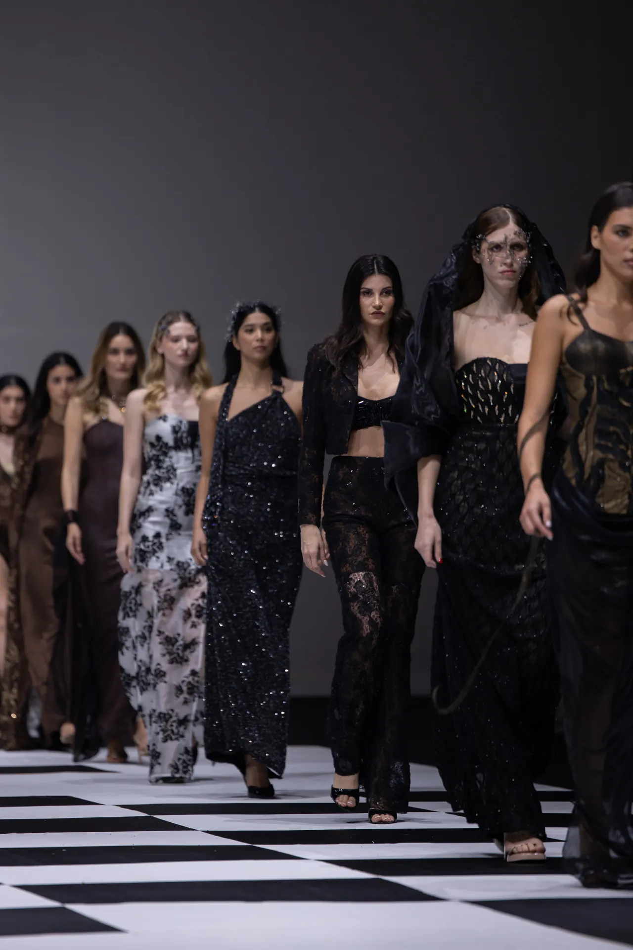 Models in dark, glittery outfits walk the fashion runway on a black-and-white checkered floor.