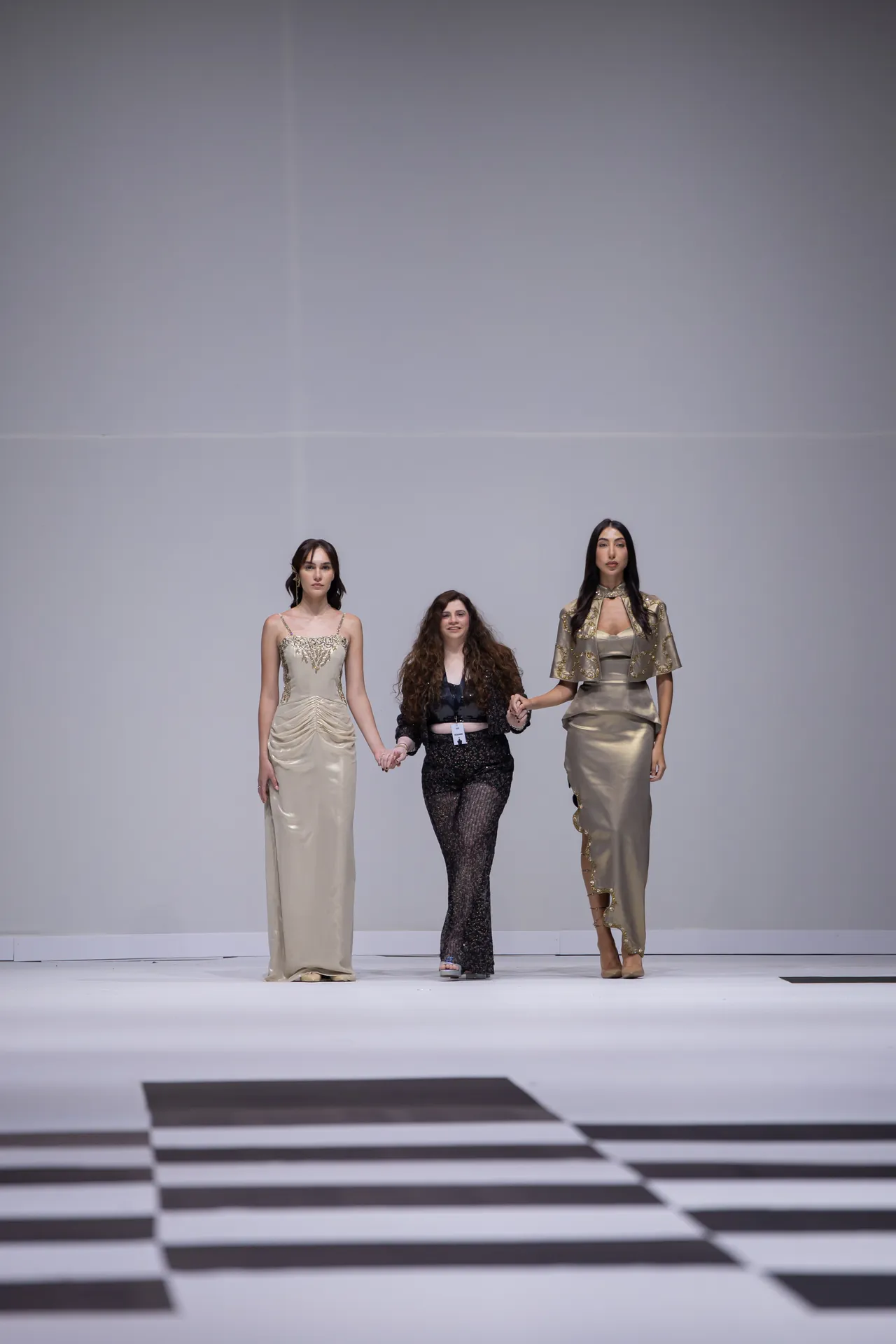 Three models walk hand-in-hand down a fashion runway, wearing elegant gold-toned gowns against a minimalist backdrop.