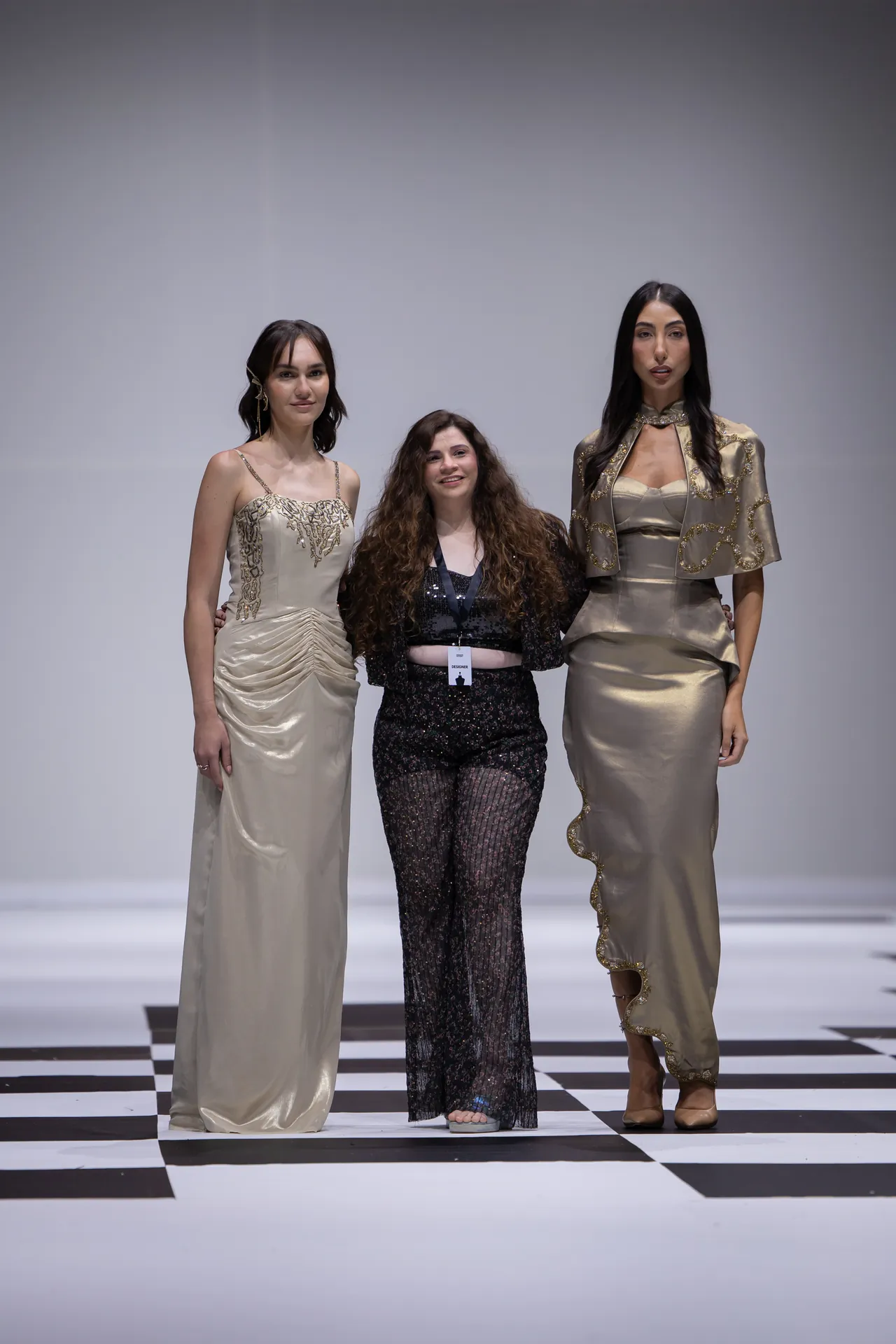 Three fashion models pose on a black-and-white checkered runway, wearing cream and gold gowns with intricate detailing.