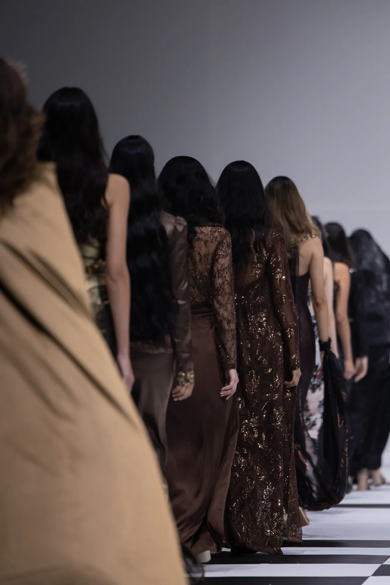 Models walk away from the camera on a fashion runway, wearing brown and black evening gowns seen from behind