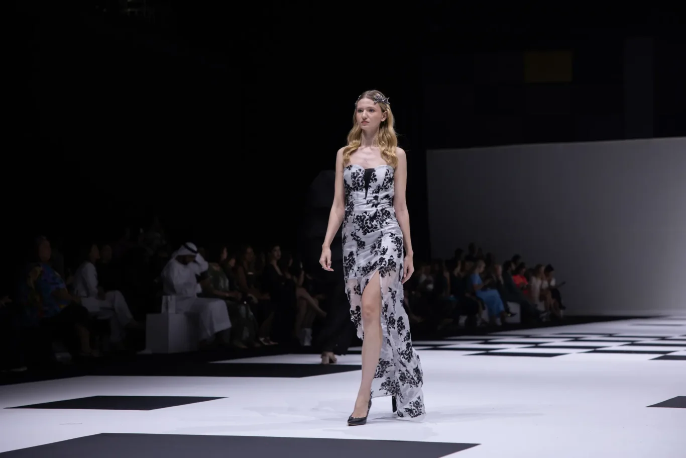 Model walks a black-and-white floral strapless dress down a white runway; audience sits in the dark in the background.