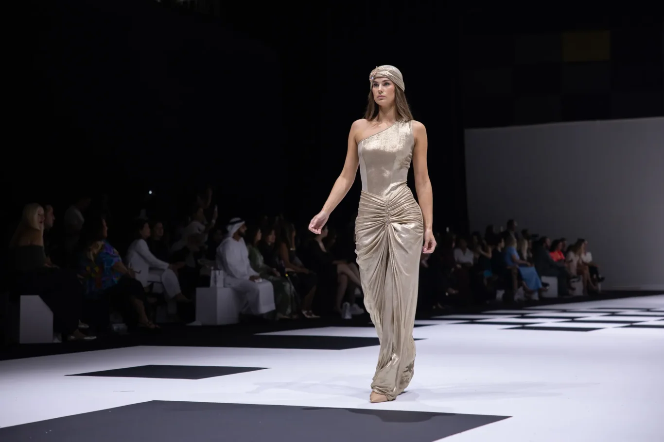 Female model walks a fashion runway in a one-shoulder metallic gold gown and headwrap, audience blurred in the background.