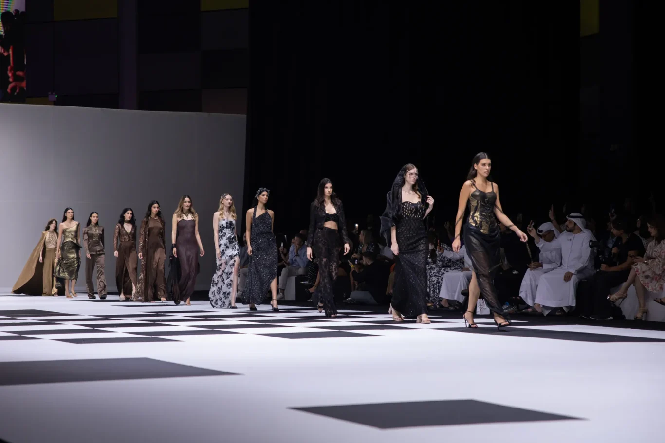 Models walk a runway in brown and black evening gowns on a black-and-white checkered floor, with spectators on the right.