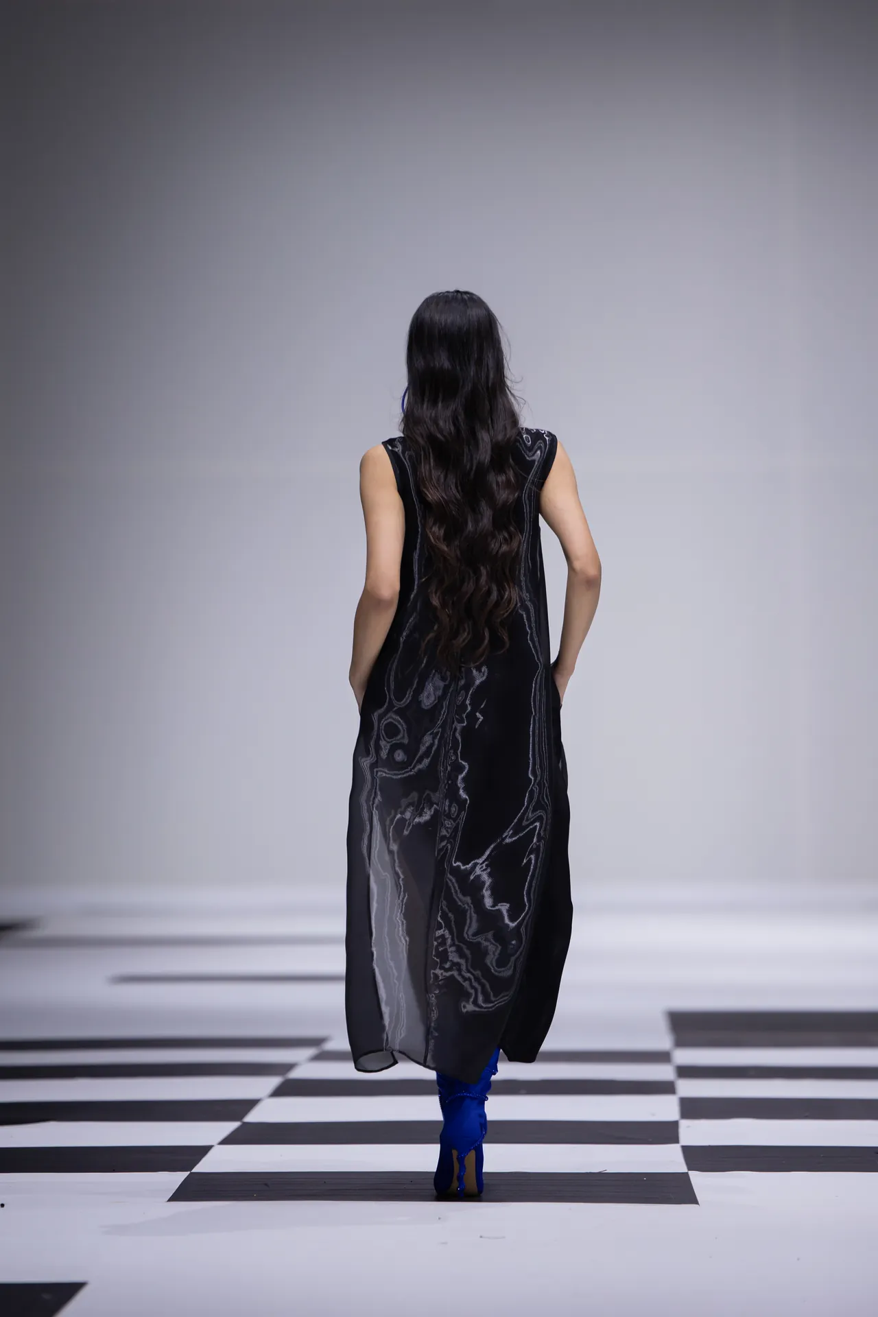 Back view of a model walking a runway in a long black patterned dress and bright blue boots on a black-and-white checkered floor.