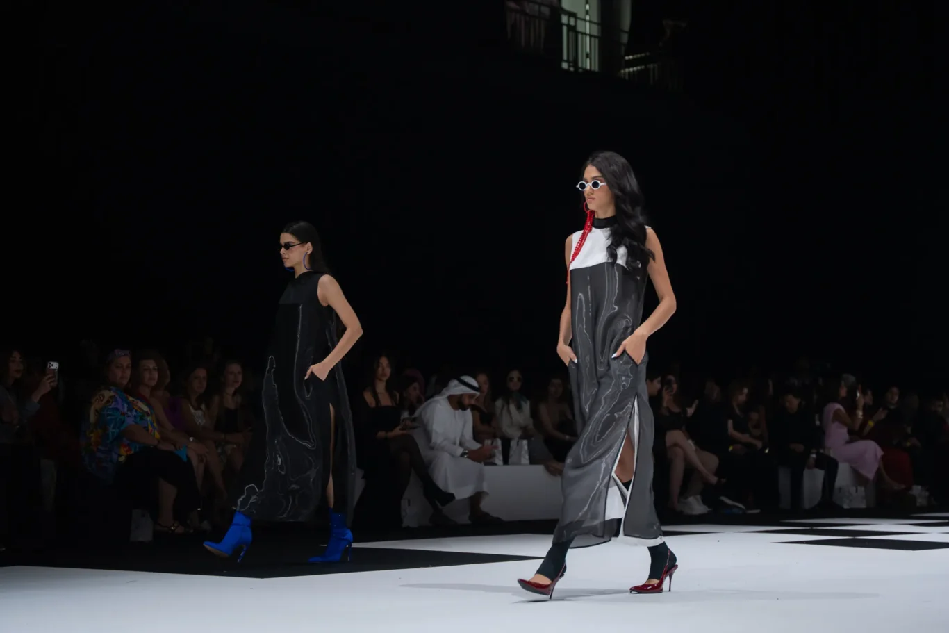 Two models walk a fashion runway in marble-pattern black and gray dresses as a seated audience watches in the dark.