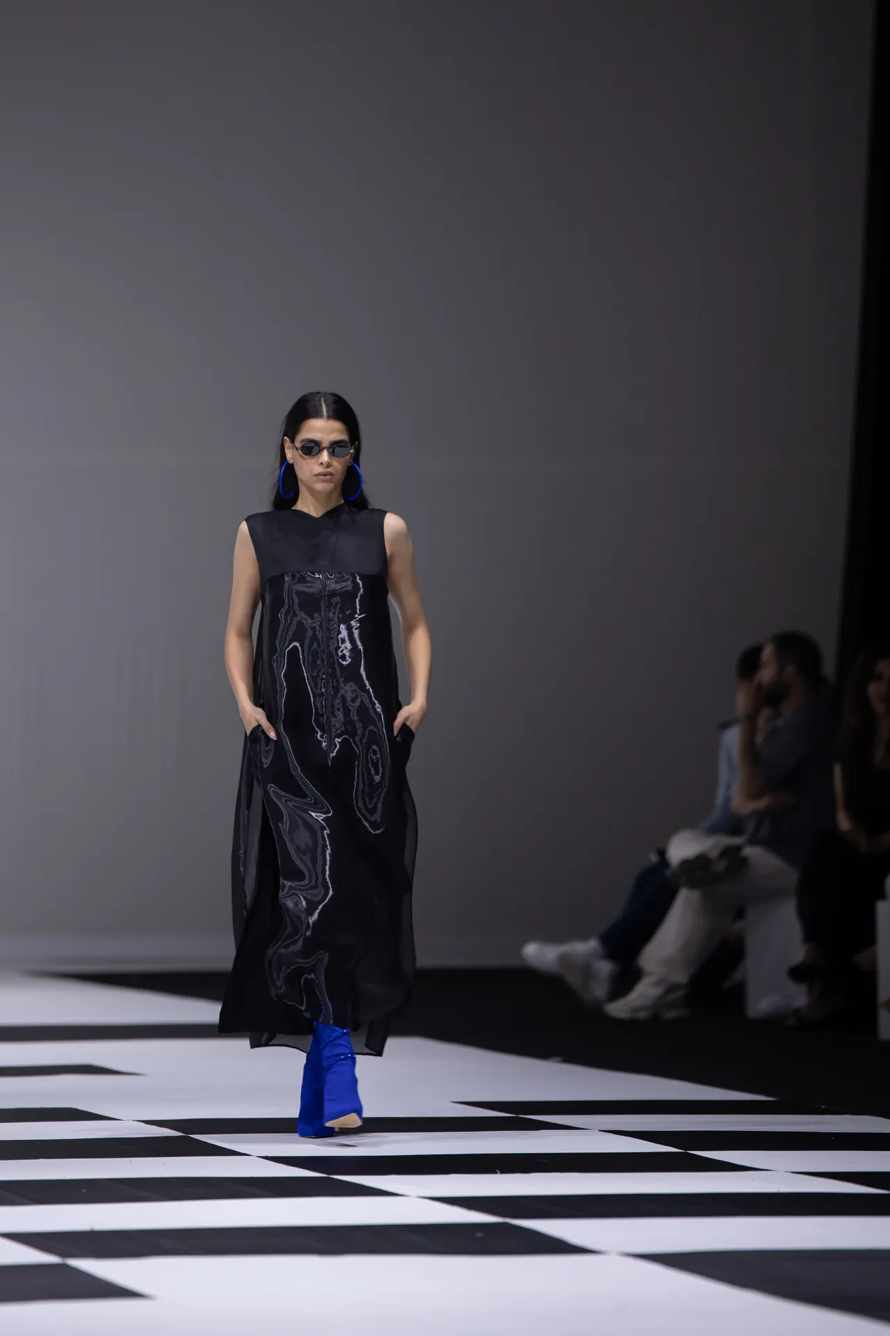 Model walks the runway in a sleeveless black dress with abstract white piping, bright blue ankle boots, blue hoop earrings, and dark sunglasses on a black-and-white checkered catwalk.