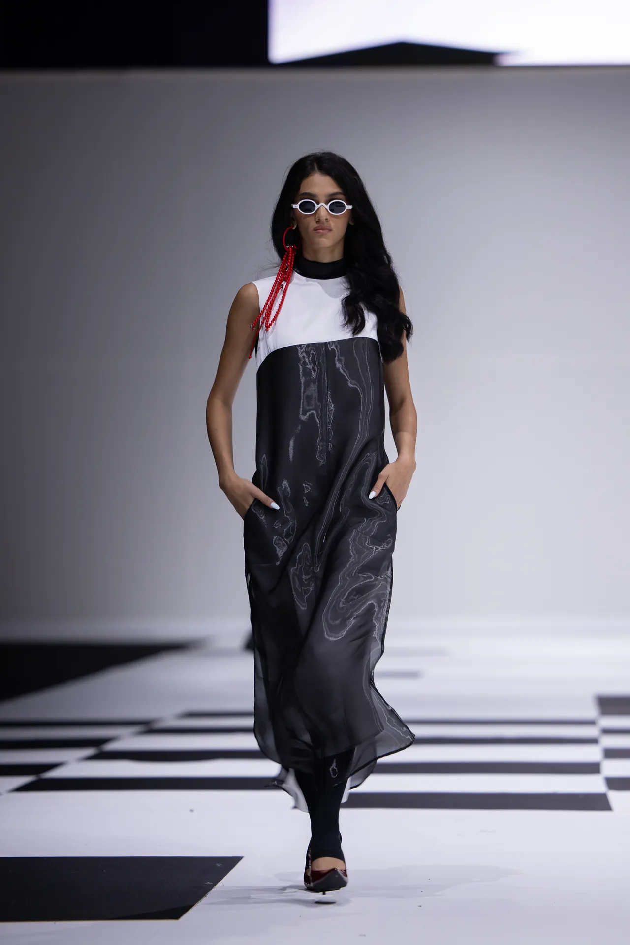 Fashion model walking a runway in a black and gray marble-print dress with a white top, red beaded earrings, and white sunglasses on a checkered catwalk.