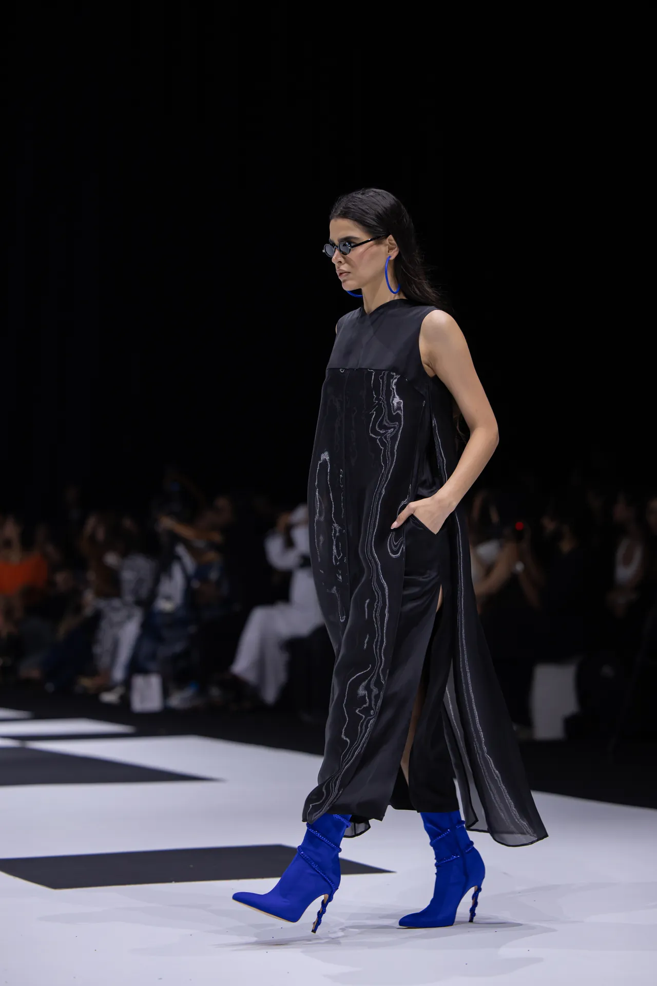 Model walking a fashion runway in a black, marbled dress with bright blue knee-high boots and matching blue hoop earrings; dark background with audience.