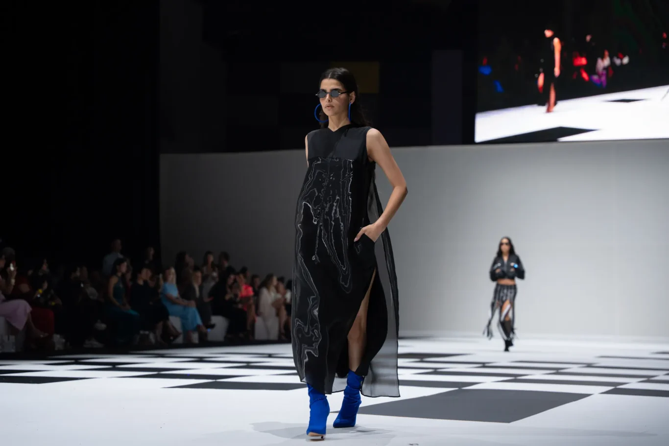 Model on a fashion runway wearing a black sleeveless dress with a white marble pattern and bright blue boots, sunglasses on.
