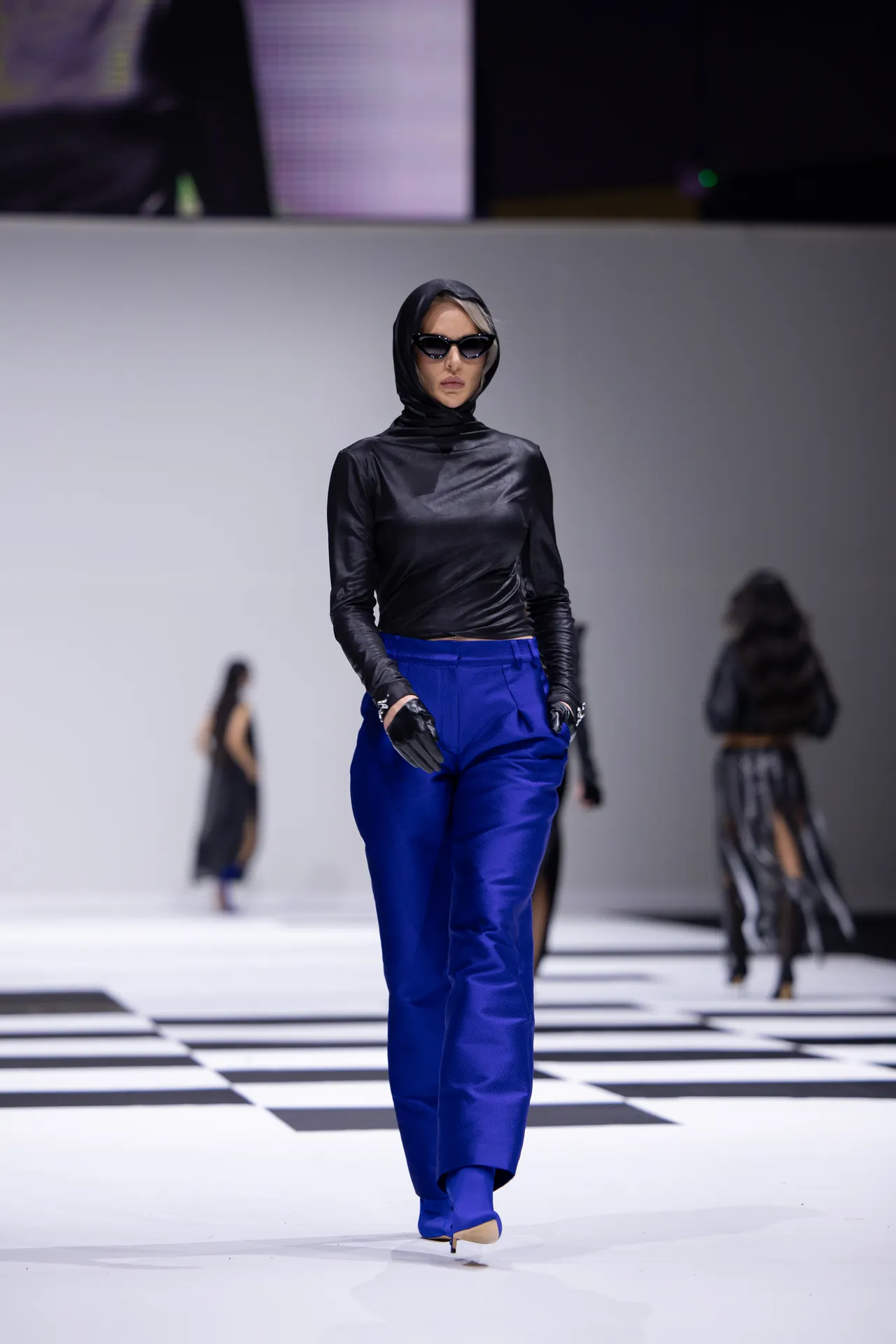Model walks runway in a black hooded top, blue wide-leg pants, and black gloves with sunglasses.