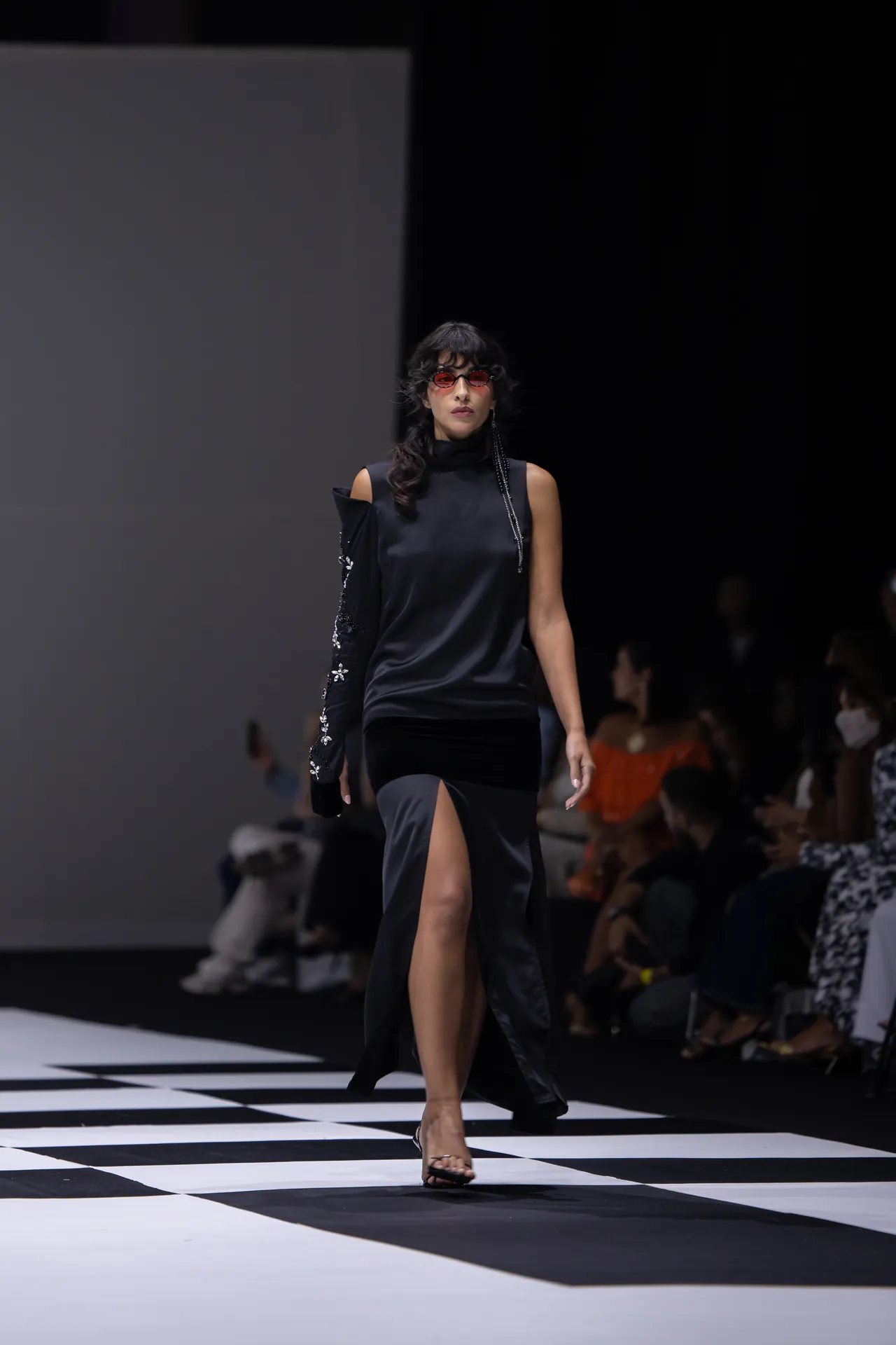 Model walking a fashion show on a black-and-white checkered runway, wearing a black sleeveless dress and red sunglasses.