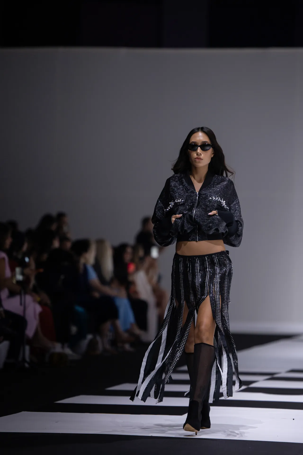 Model walks a fashion runway in a black, glittery jacket and matching fringe skirt with sunglasses, audience in the background.