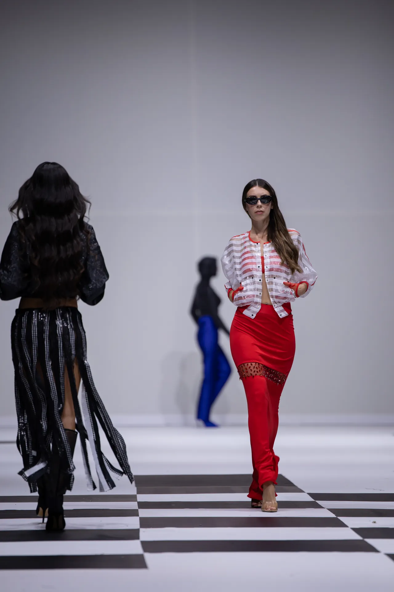 Model in red pants and a striped sheer jacket walks the runway on a black-and-white checkerboard floor, wearing sunglasses and heels.