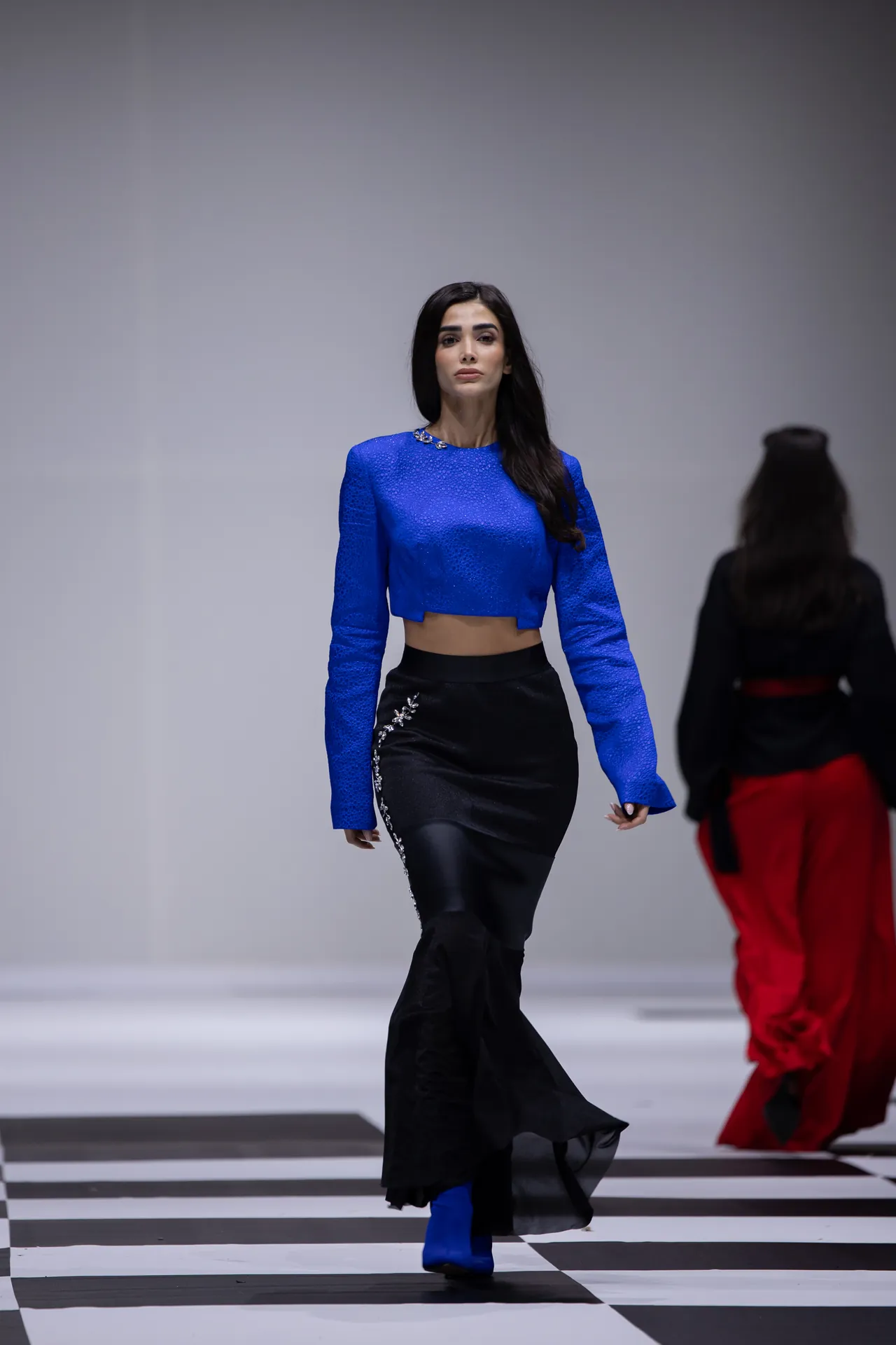 Model walking a fashion runway in a bright blue cropped top and black embellished skirt, blue boots, on a black-and-white striped floor.