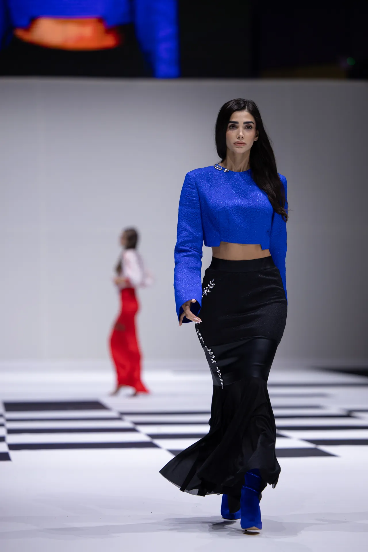 Model walks a runway in a royal blue cropped top and black embellished mermaid skirt amid a black-and-white checker floor.