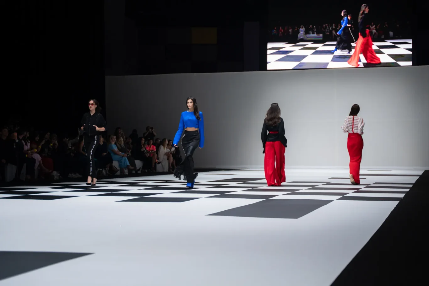 Models walk a black-and-white checkered runway during a fashion show with spectators seated along the left. A large screen above shows more runway footage. A blue-clad model strides center stage, flanked by other models in red pants and black outfits.