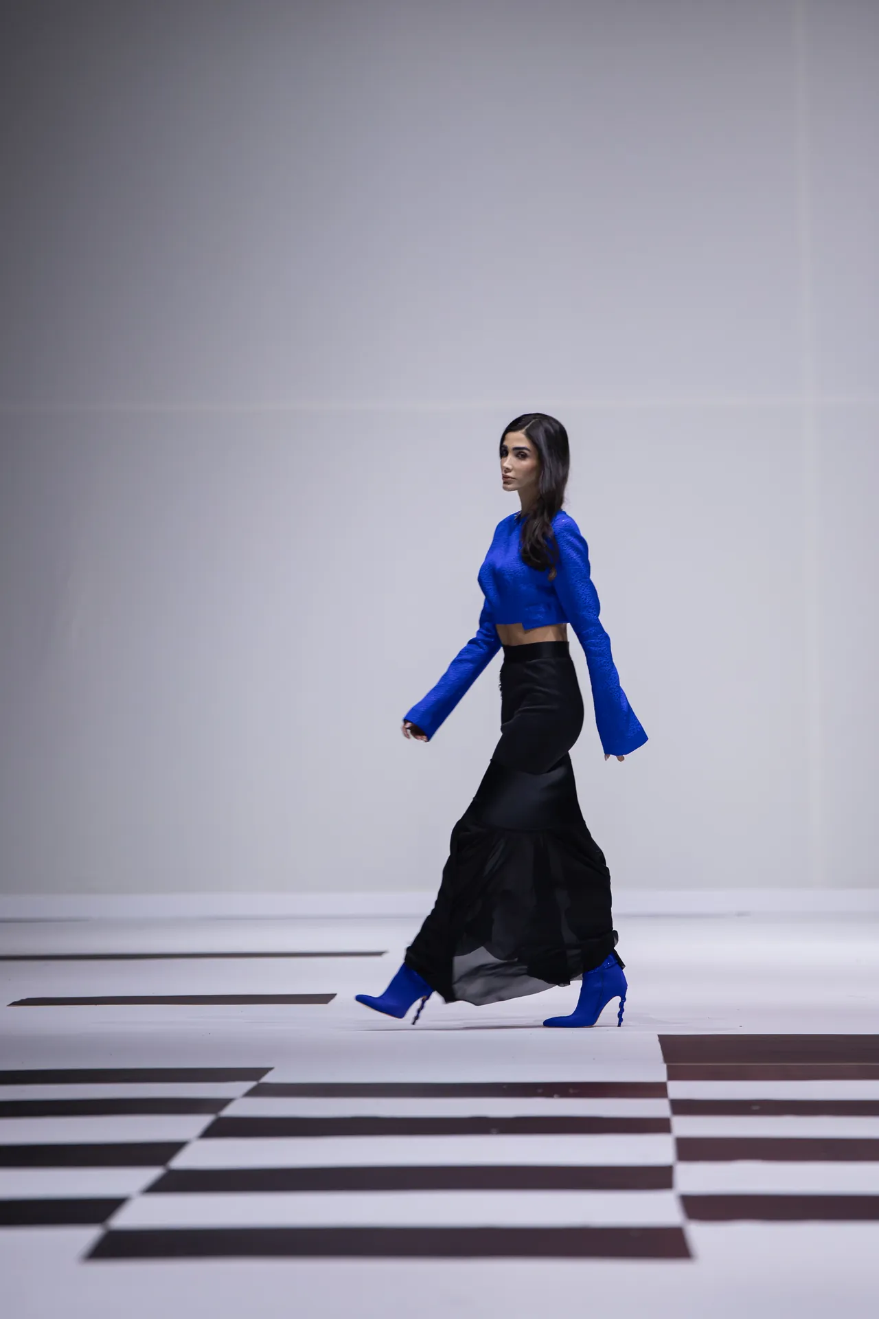 Model walking on a white runway in a blue cropped top, black skirt, and blue boots, against a minimalist backdrop.