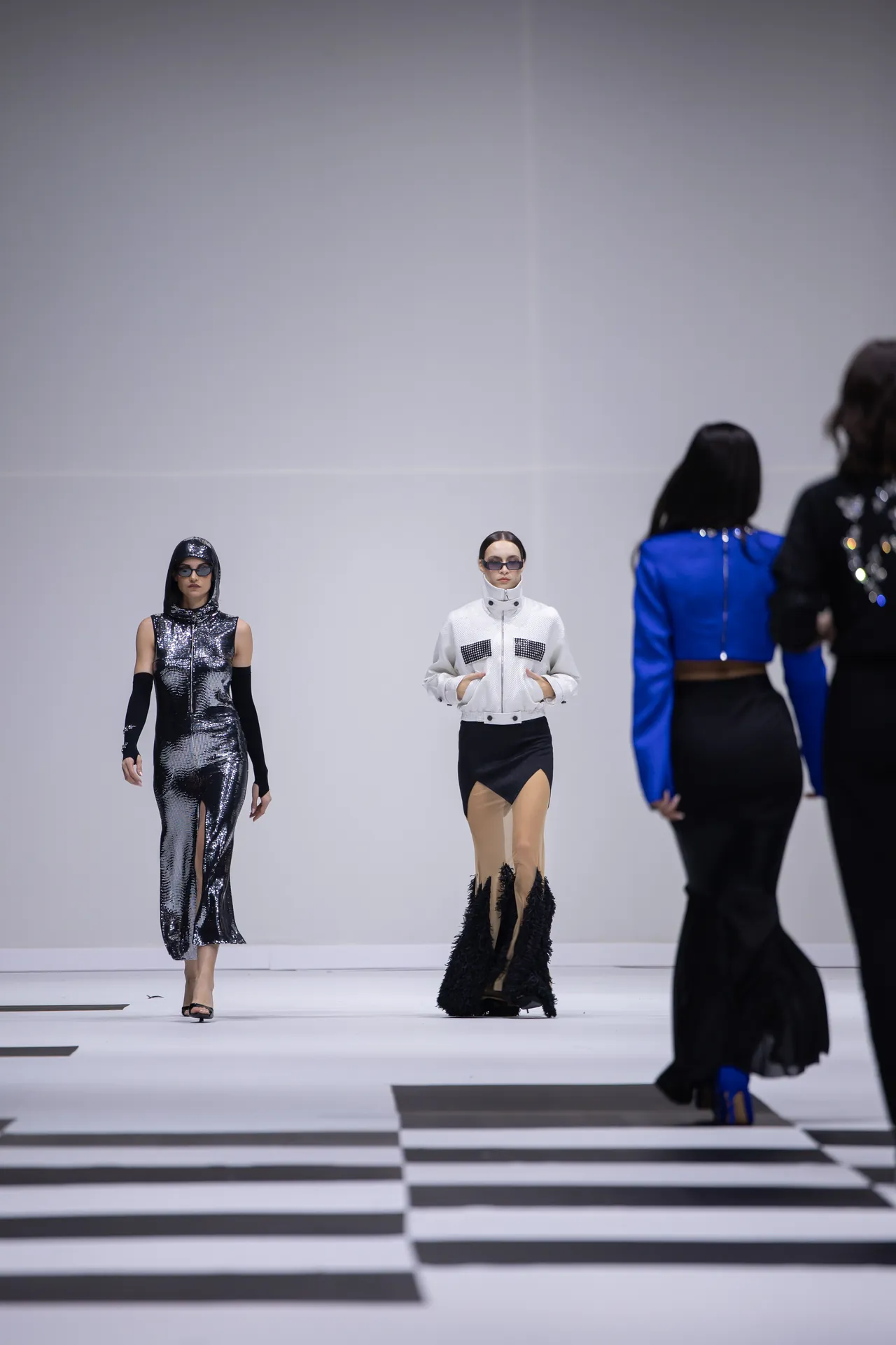 Models walk a minimalist white runway with a black-and-white striped floor, wearing futuristic metallic outfits.