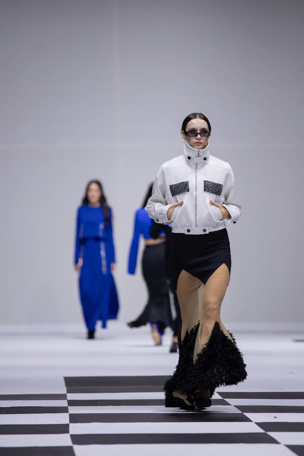 Model walking a fashion runway in a white textured jacket with studded pockets and a black feather-trim skirt; blue-clad models visible in the blurred background on a checkered floor.