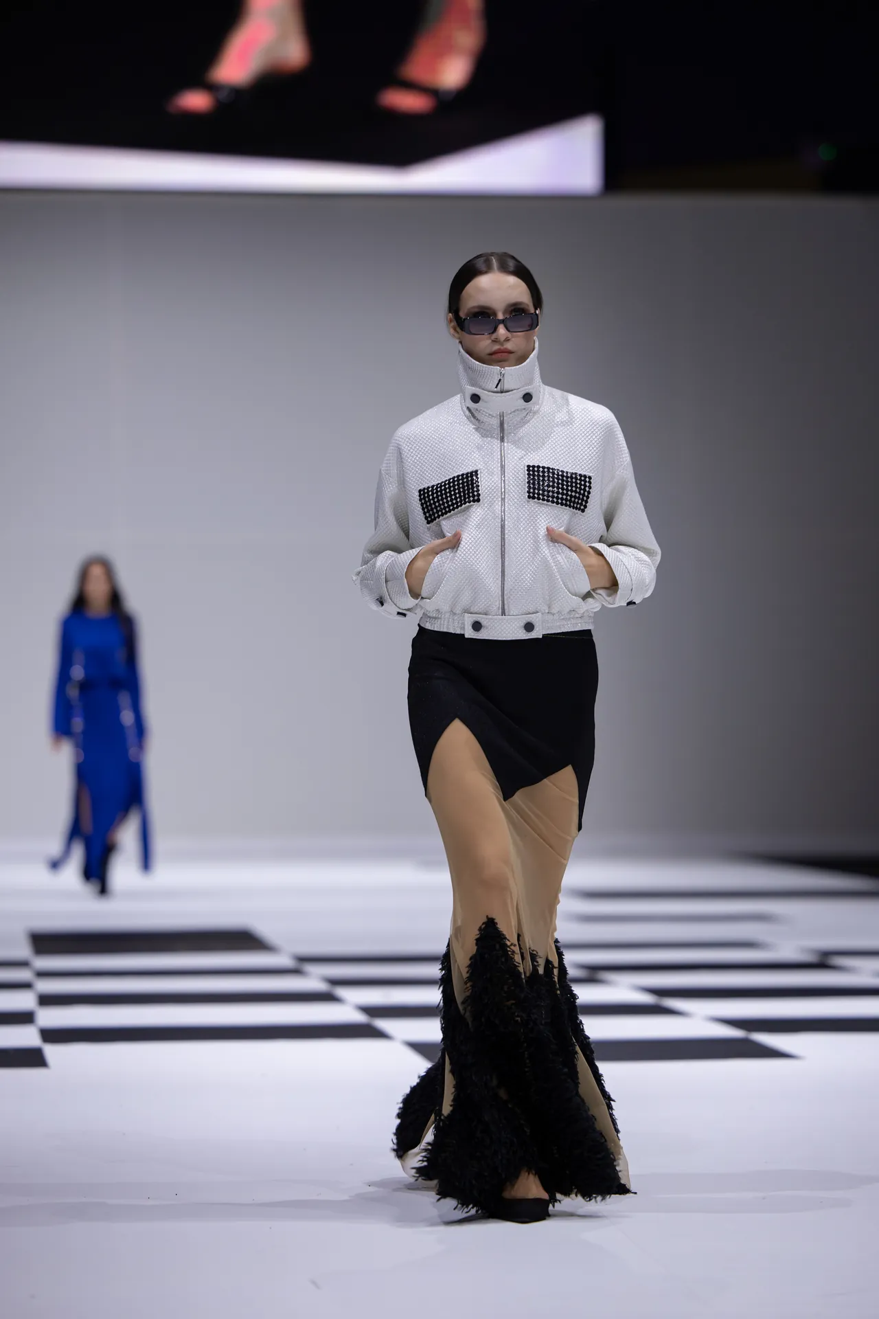 Model on a fashion runway wearing a white textured jacket with studded pockets and a black asymmetric skirt with a feathered hem, walking on a black-and-white checkered stage.