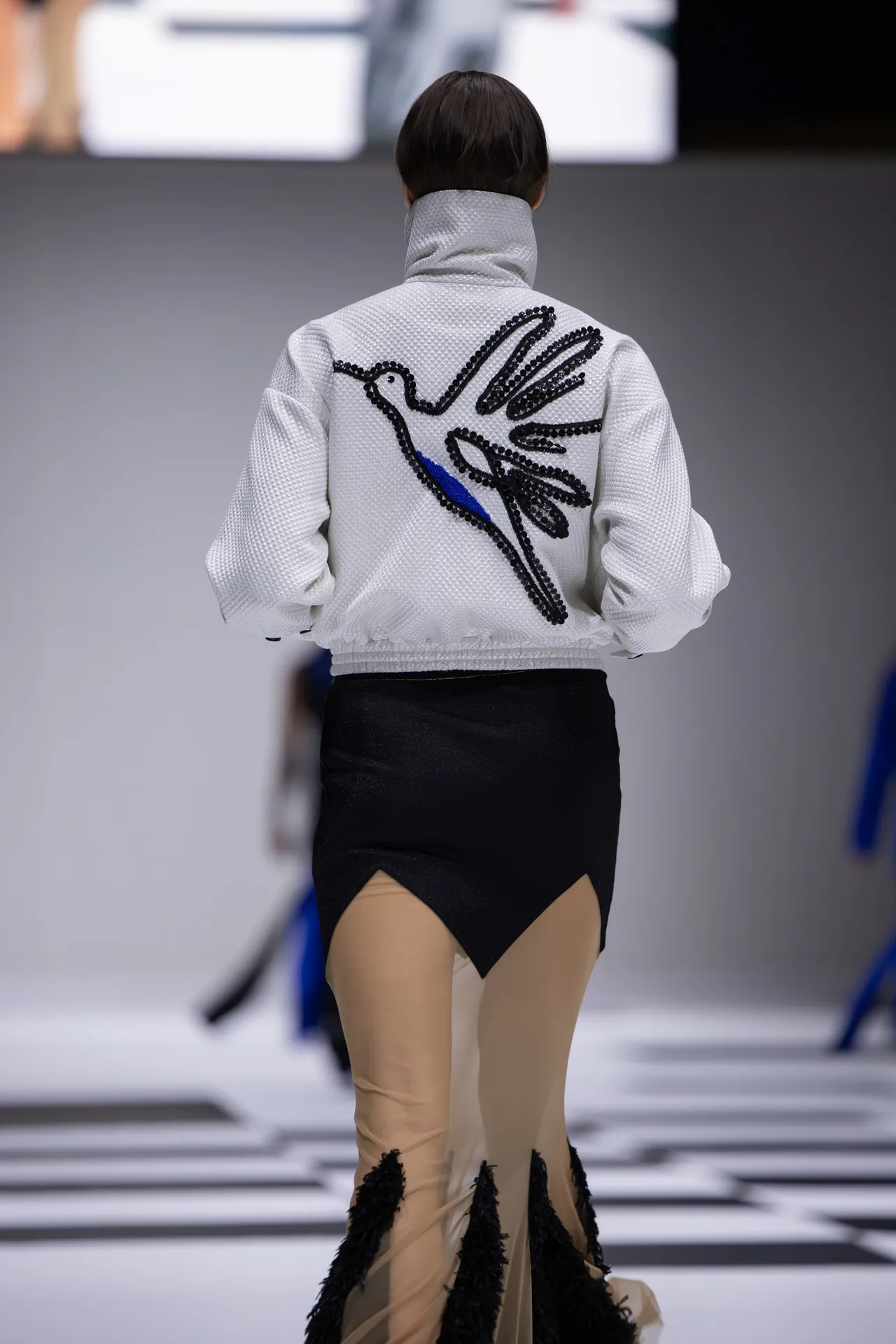 Back view of a fashion model wearing a white textured jacket with a black sequin bird design and a black asymmetrical skirt on a runway.