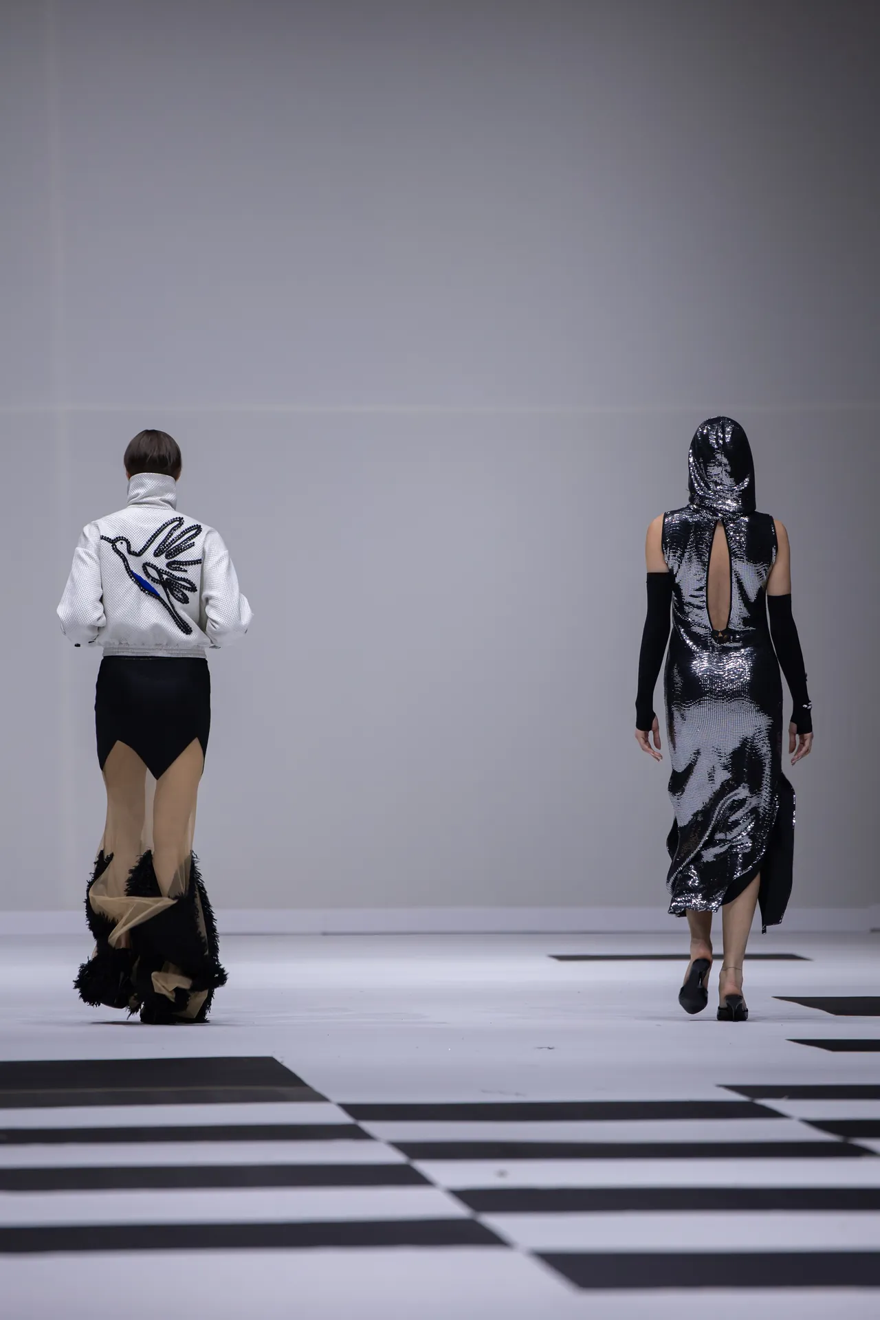 Two models walk a fashion runway on a black-and-white striped floor; one wears a white jacket with a black skirt and fringe, the other a metallic silver hooded dress with open back details.