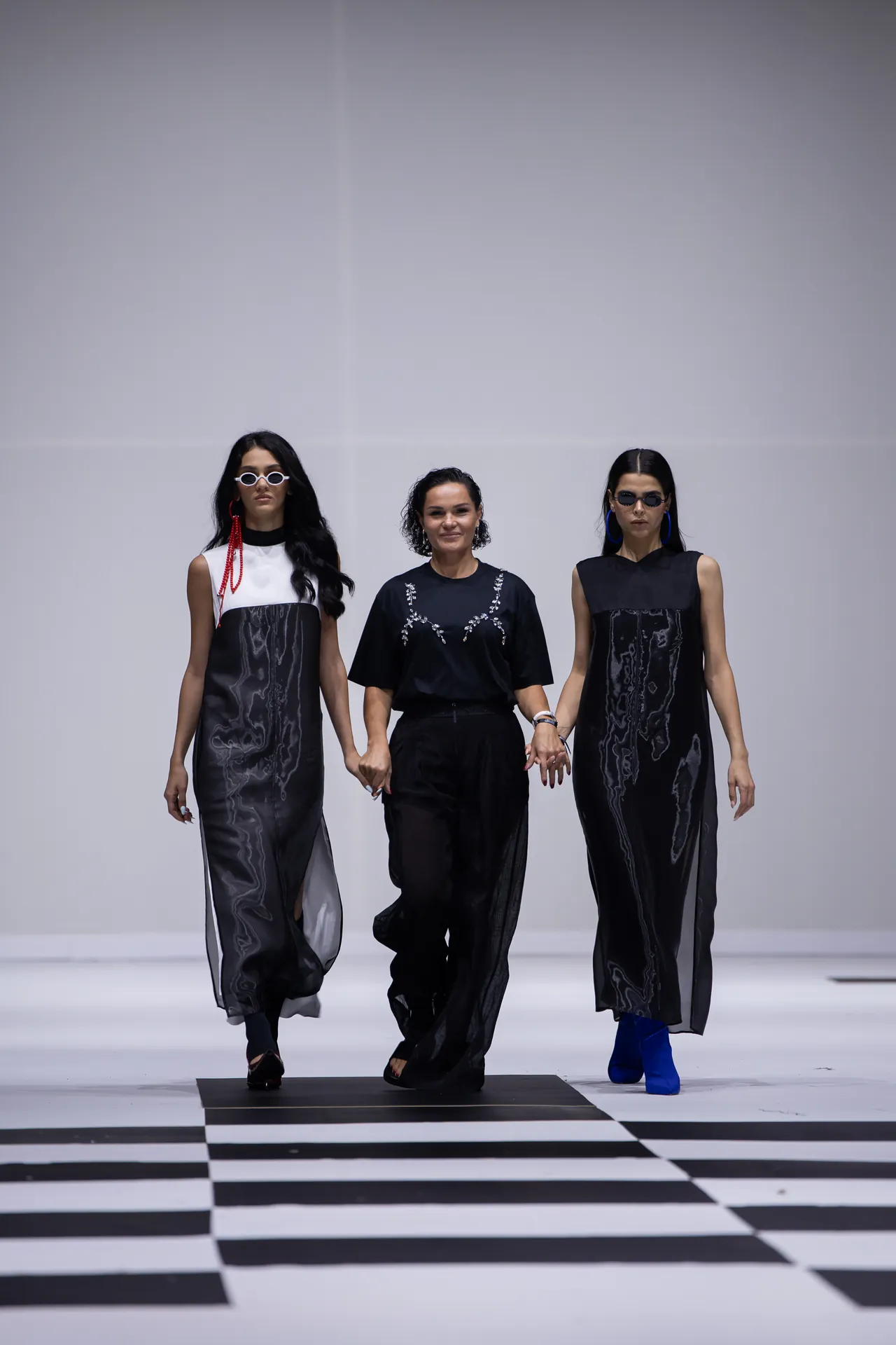 Three fashion models walk hand in hand down a runway in black outfits against a white minimalist backdrop.