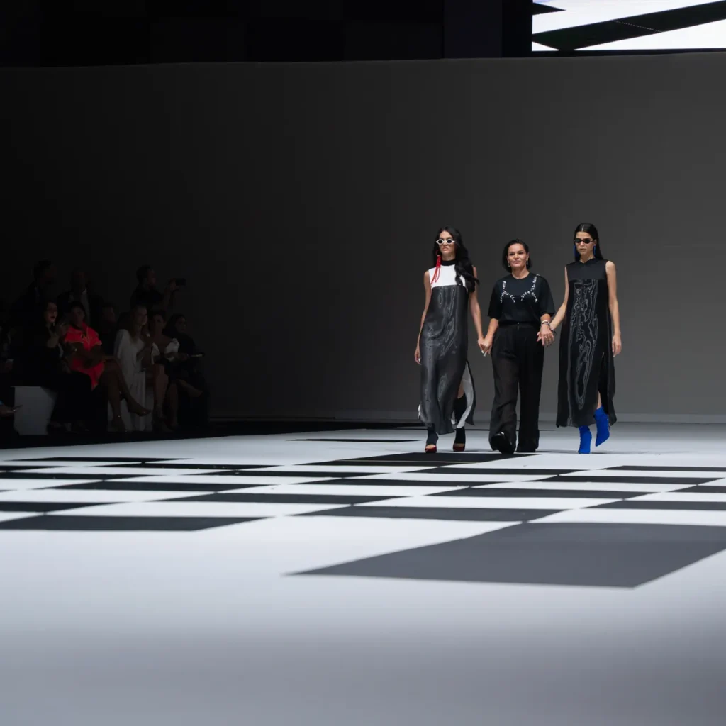 Three models walk a fashion runway holding hands, wearing black outfits with silver patterns on a black-and-white geometric floor, audience along the left wall.
