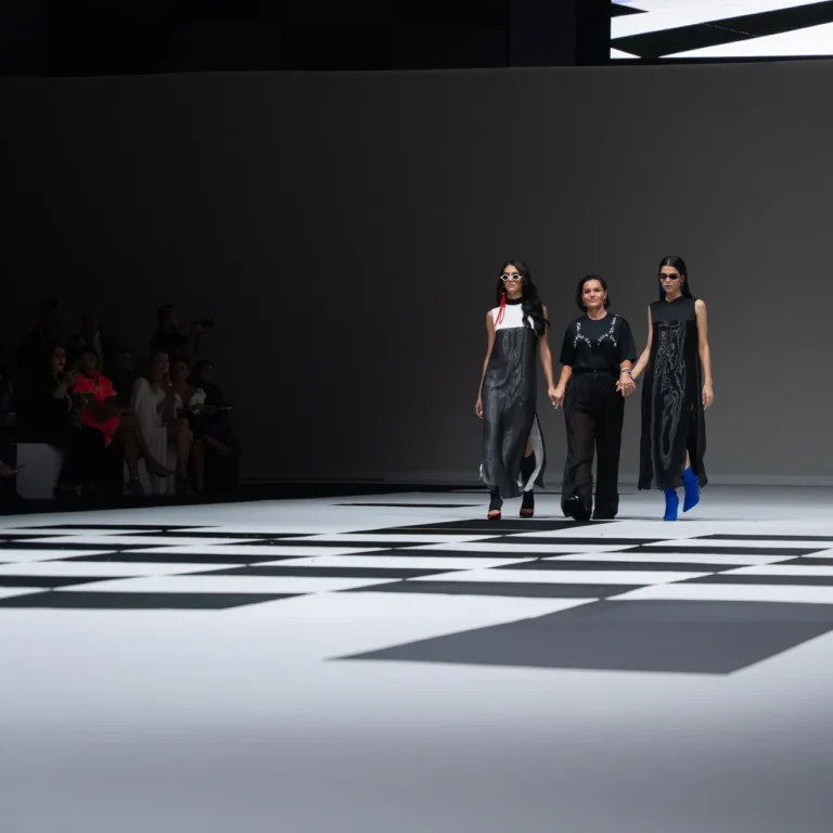 Three models walk a fashion runway holding hands, wearing black outfits with silver patterns on a black-and-white geometric floor, audience along the left wall.