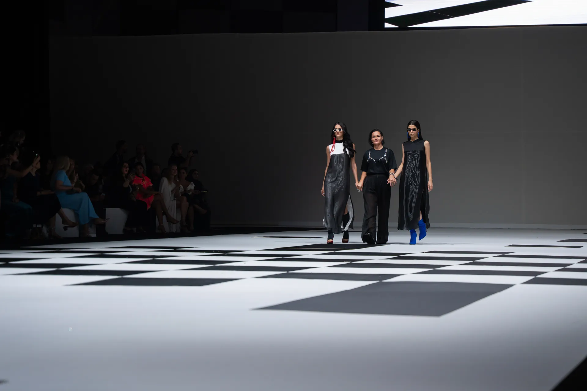 Three models walk a fashion runway holding hands, wearing black outfits with silver patterns on a black-and-white geometric floor, audience along the left wall.