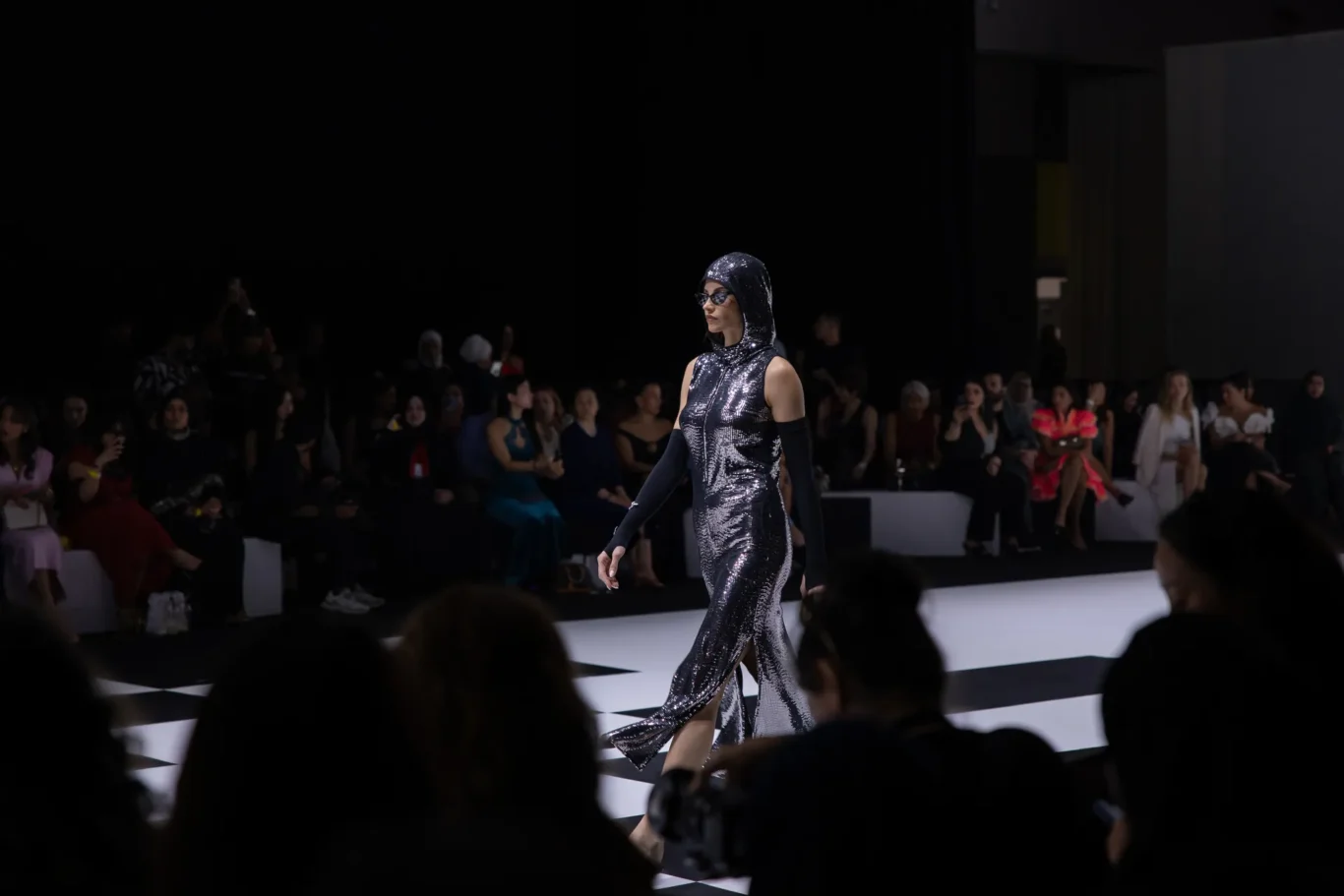 Model in a silver sequined hooded dress walks the runway; audience sits in dim lighting in the background.