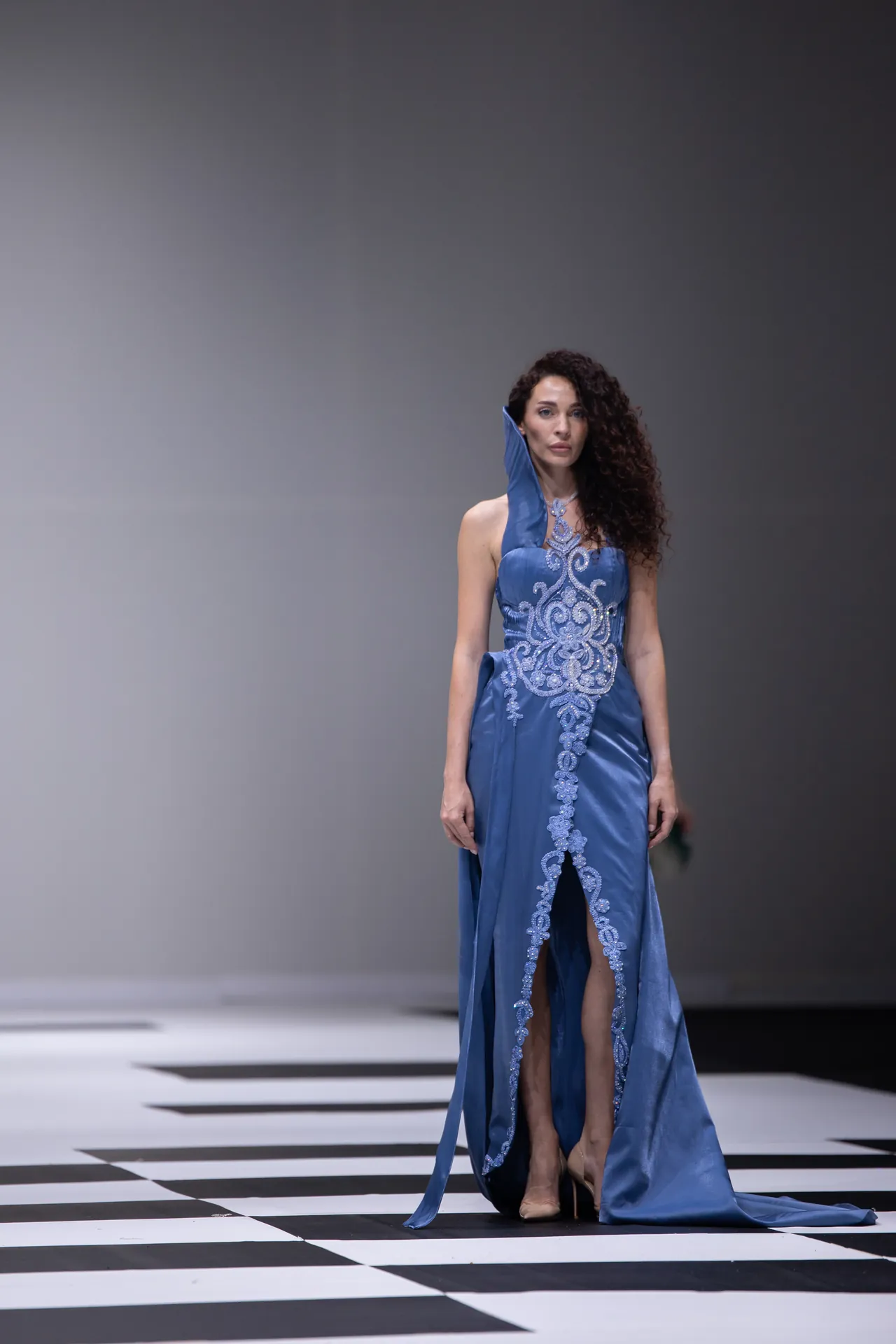 Model in a blue, intricately embroidered evening gown on a black-and-white checkered runway