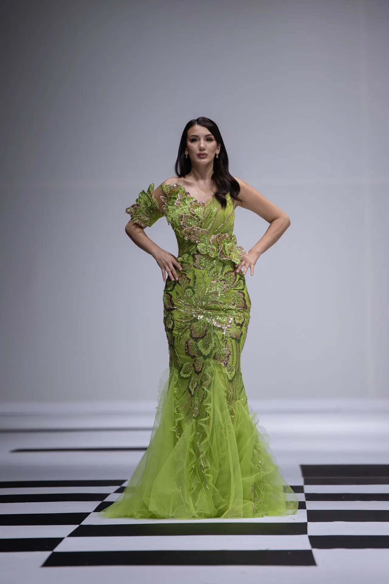 Model on a runway wearing a lime green, embellished gown with floral appliqués and sheer train, posing with hands on hips on a checkered floor.