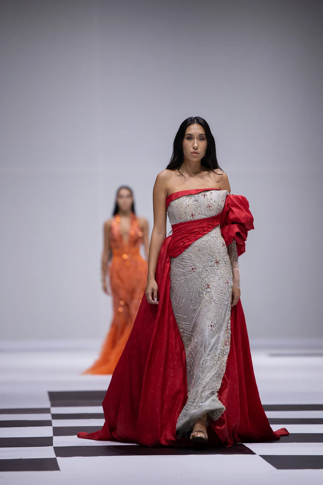 Model walks a fashion runway in a silver beaded gown with a red sash and flowing red cape; a second model in an orange dress appears blurred in the background on a black-and-white checkered floor.